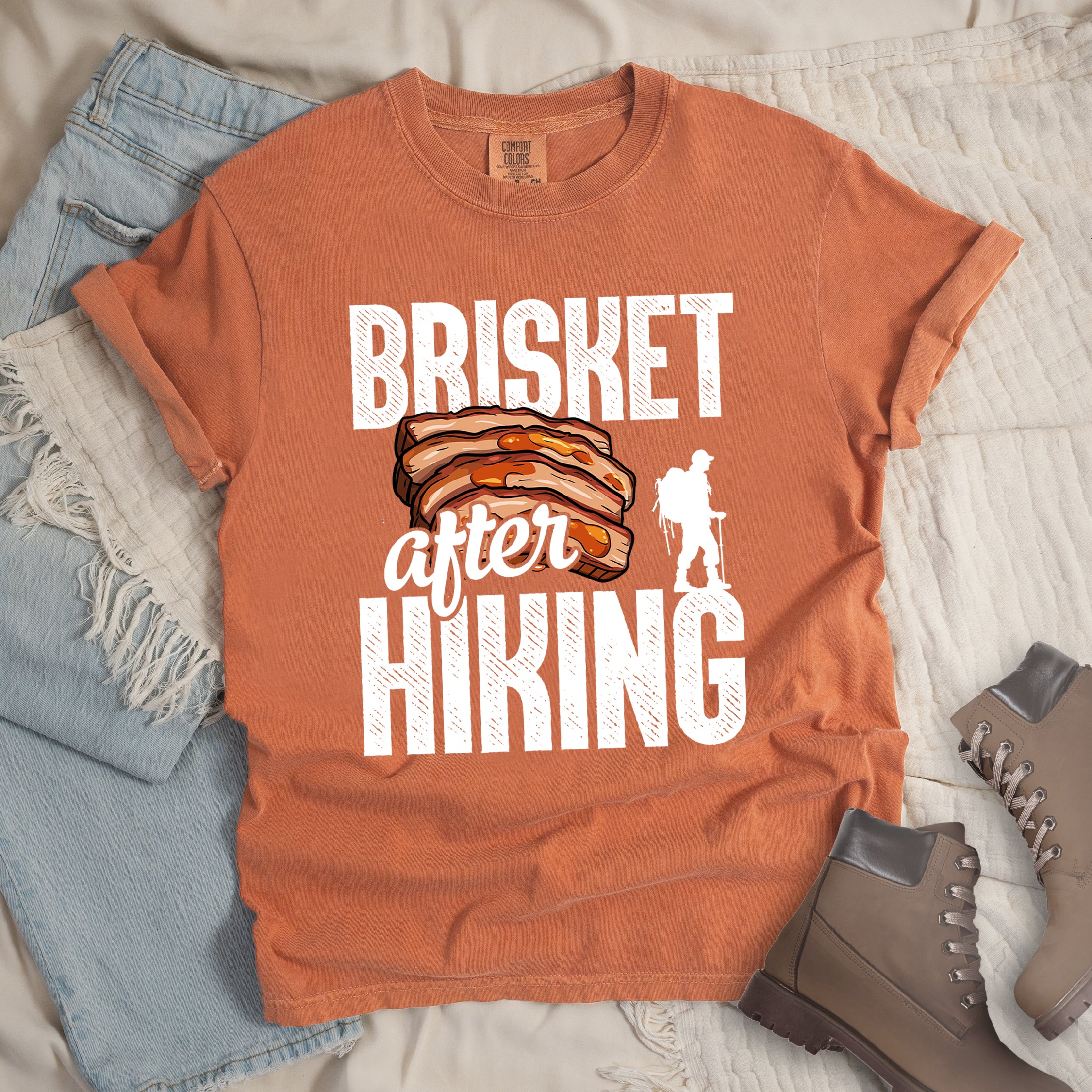 Yam (Brown) t-shirt with white distressed text reading "BRISKET after HIKING". "BRISKET" appears in large white letters at the top, "after" in white script in the middle, and "HIKING" in large white letters at the bottom. Features a detailed illustration of sliced brisket meat in orange-brown tones in the center and a white silhouette of a hiker with walking poles on the right side.