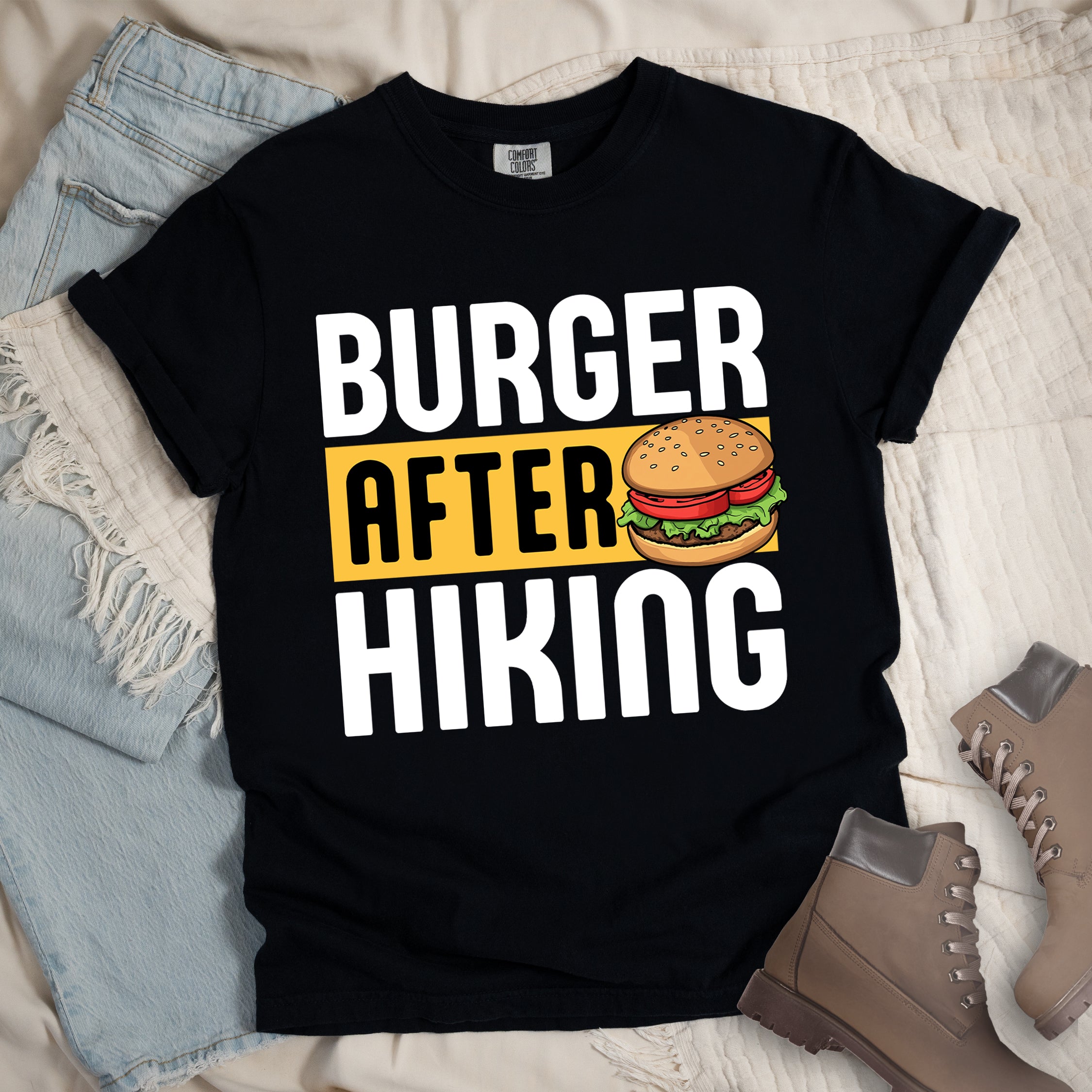 Black t-shirt with bold text reading "BURGER AFTER HIKING". "BURGER" appears in large white letters at the top, "AFTER" in black text on a yellow rectangular background in the middle, and "HIKING" in large white letters at the bottom. Features a detailed illustration of a hamburger with sesame seed bun, lettuce, tomato and patty next to the word "AFTER".  The shirt features a classic crew neck and casual rolled sleeves, styled alongside light wash denim and brown hiking boots.