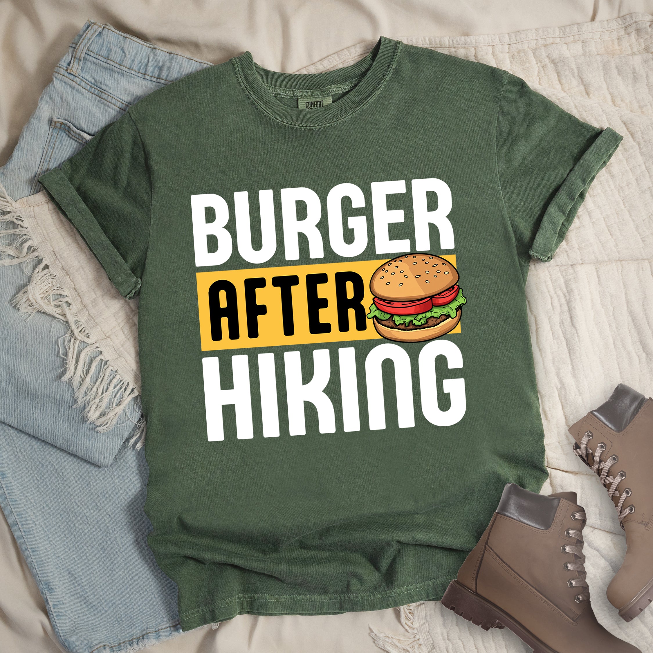 Moss (Dark Green) t-shirt with bold text reading "BURGER AFTER HIKING". "BURGER" appears in large white letters at the top, "AFTER" in black text on a yellow rectangular background in the middle, and "HIKING" in large white letters at the bottom. Features a detailed illustration of a hamburger with sesame seed bun, lettuce, tomato and patty next to the word "AFTER".  The shirt features a classic crew neck and casual rolled sleeves, styled alongside light wash denim and brown hiking boots.
