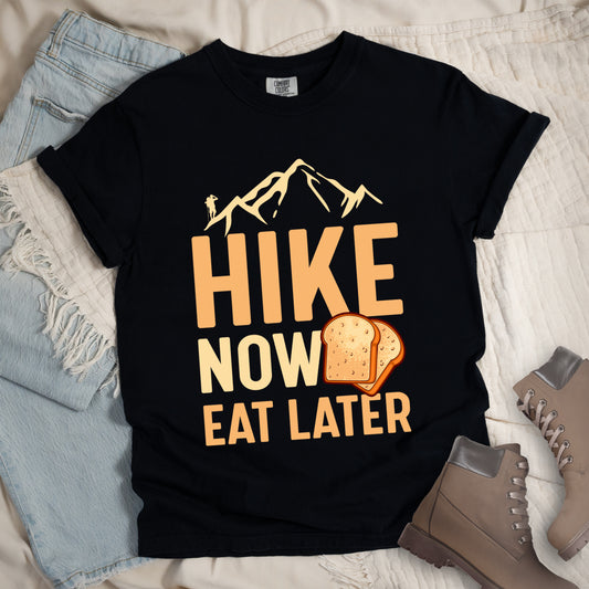 A black t-shirt with a design showing "HIKE NOW EAT LATER" in gradient orange and tan text. Above "HIKE" is a mountain silhouette with a small hiker figure. A sandwich/toast illustration appears between "NOW" and "EAT LATER". The text is arranged in three lines with "HIKE" at the top in largest font, followed by "NOW" and "EAT LATER" below. The shirt features a classic crew neck and casual rolled sleeves, styled alongside light wash denim and brown hiking boots.