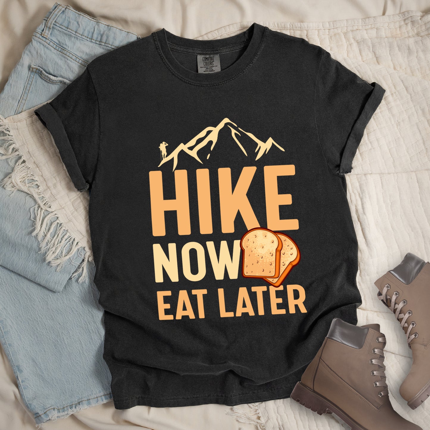 A Graphite (Deep Gray) t-shirt with a design showing "HIKE NOW EAT LATER" in gradient orange and tan text. Above "HIKE" is a mountain silhouette with a small hiker figure. A sandwich/toast illustration appears between "NOW" and "EAT LATER". The text is arranged in three lines with "HIKE" at the top in largest font, followed by "NOW" and "EAT LATER" below. The shirt features a classic crew neck and casual rolled sleeves, styled alongside light wash denim and brown hiking boots.