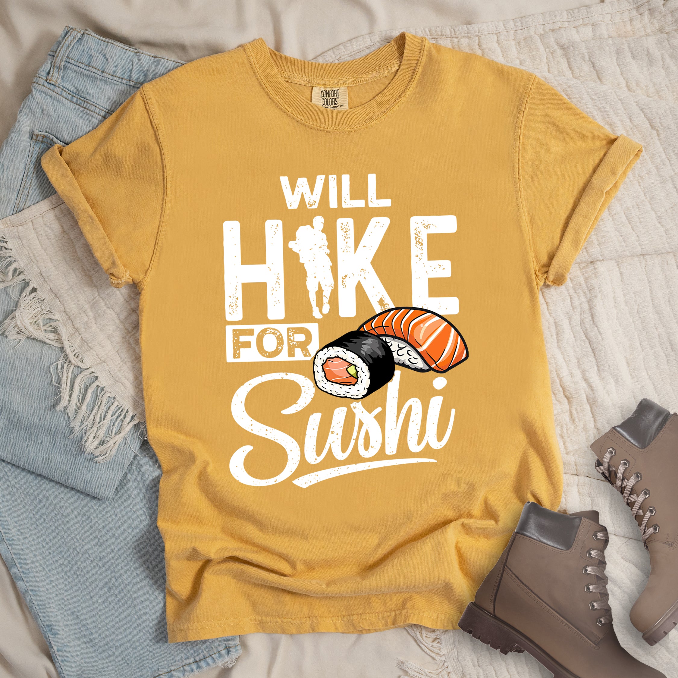 Mustard (Yellow) t-shirt with white distressed text reading "WILL HIKE FOR Sushi". "WILL" appears at the top, "HIKE" in large letters with a hiker silhouette integrated into the "I", "FOR" in smaller text, and "Sushi" in stylized script at the bottom. Features colorful sushi illustrations including a salmon nigiri and a maki roll. Features a classic crew neck and casual rolled sleeves, styled alongside light wash denim and brown hiking boots. 