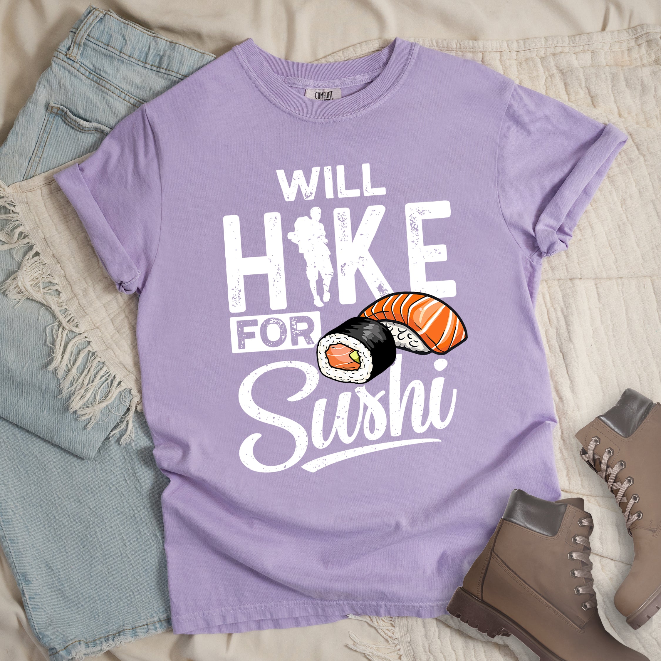 Orchid (Light Violet) t-shirt with white distressed text reading "WILL HIKE FOR Sushi". "WILL" appears at the top, "HIKE" in large letters with a hiker silhouette integrated into the "I", "FOR" in smaller text, and "Sushi" in stylized script at the bottom. Features colorful sushi illustrations including a salmon nigiri and a maki roll. Features a classic crew neck and casual rolled sleeves, styled alongside light wash denim and brown hiking boots. 