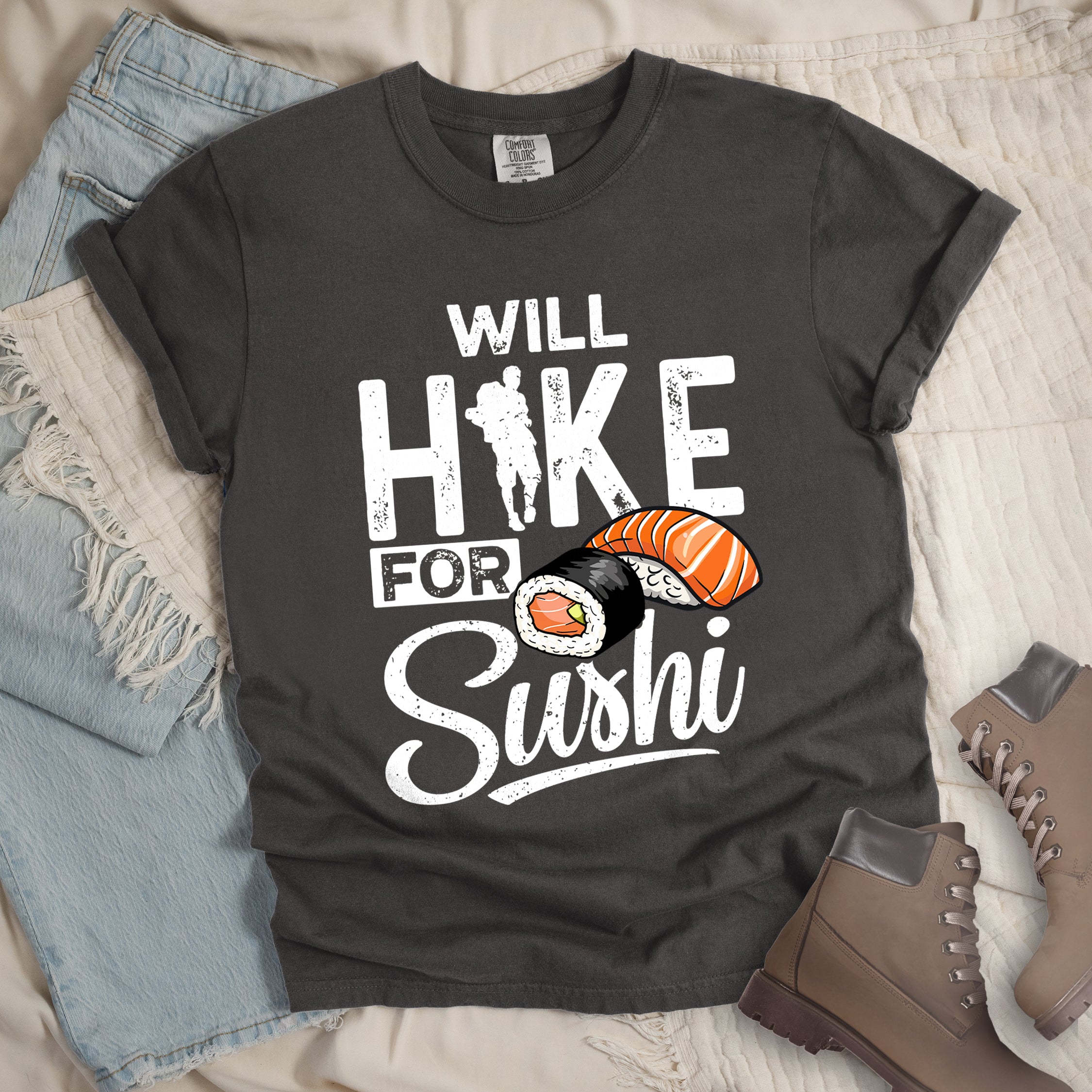 Pepper (Dark Gray) t-shirt with white distressed text reading "WILL HIKE FOR Sushi". "WILL" appears at the top, "HIKE" in large letters with a hiker silhouette integrated into the "I", "FOR" in smaller text, and "Sushi" in stylized script at the bottom. Features colorful sushi illustrations including a salmon nigiri and a maki roll. Features a classic crew neck and casual rolled sleeves, styled alongside light wash denim and brown hiking boots. 