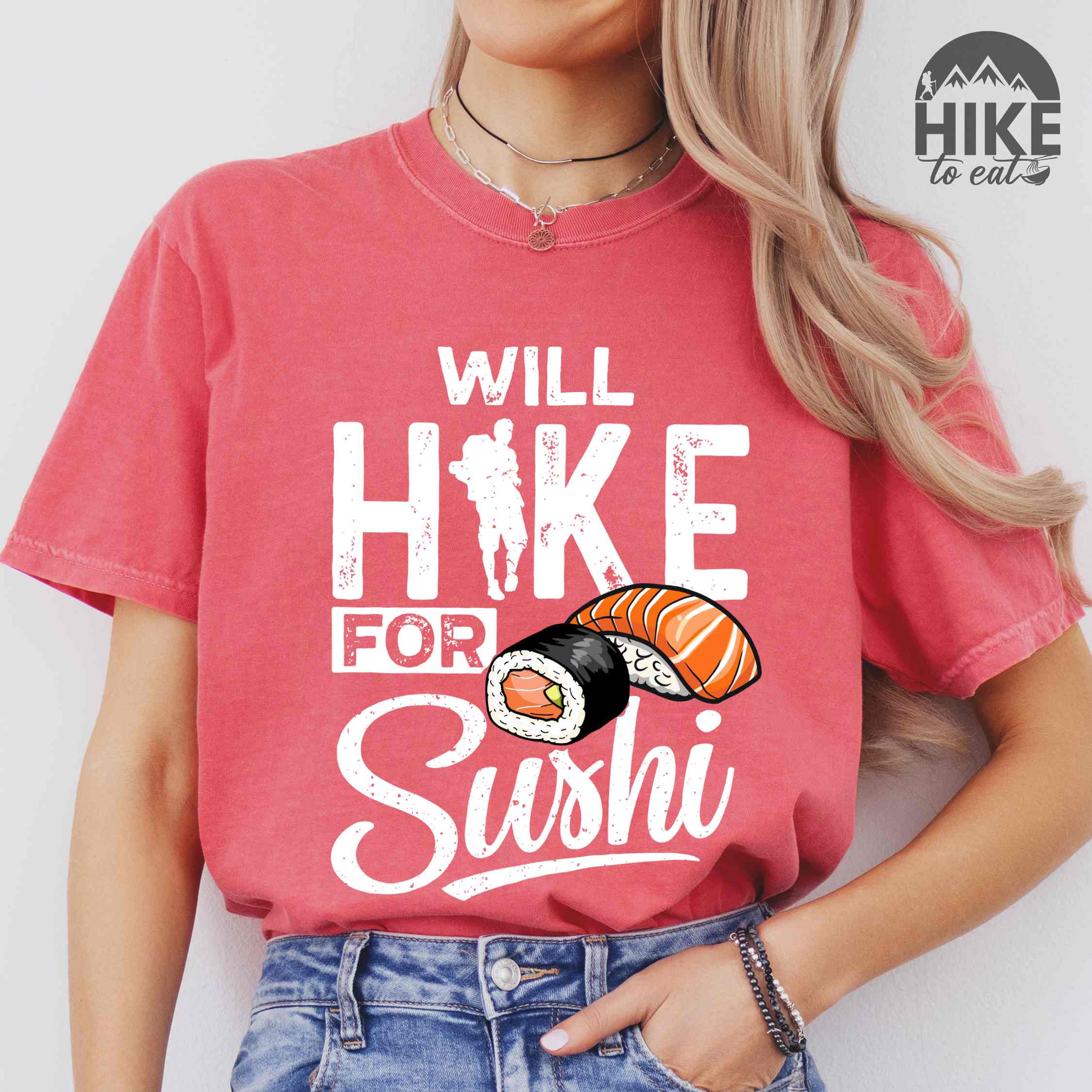 Woman wearing watermelon "Will hike for sushi" colored vintage tee with bold typography, birds, mountains, illustrated sushi platter, and backpacker for sushi lovers.
