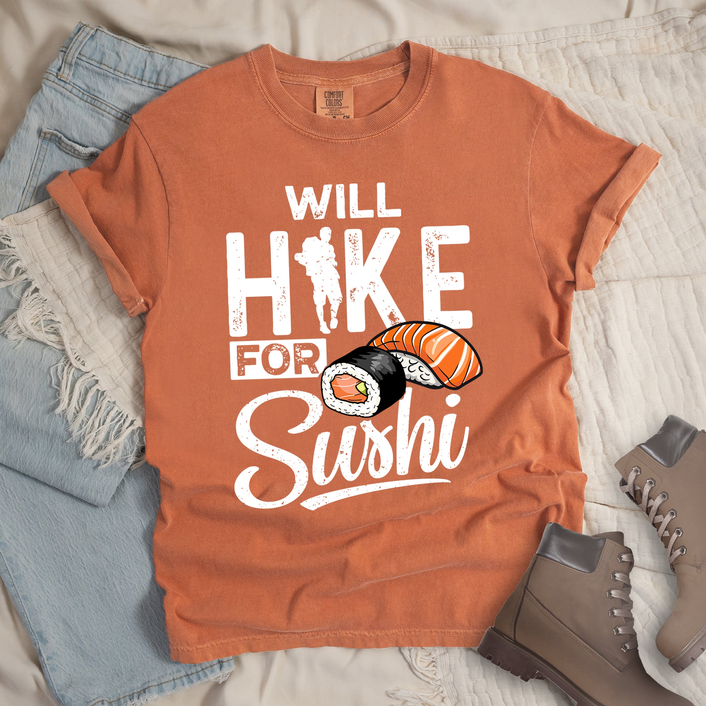 Yam (Brown) t-shirt with white distressed text reading "WILL HIKE FOR Sushi". "WILL" appears at the top, "HIKE" in large letters with a hiker silhouette integrated into the "I", "FOR" in smaller text, and "Sushi" in stylized script at the bottom. Features colorful sushi illustrations including a salmon nigiri and a maki roll. Features a classic crew neck and casual rolled sleeves, styled alongside light wash denim and brown hiking boots. 