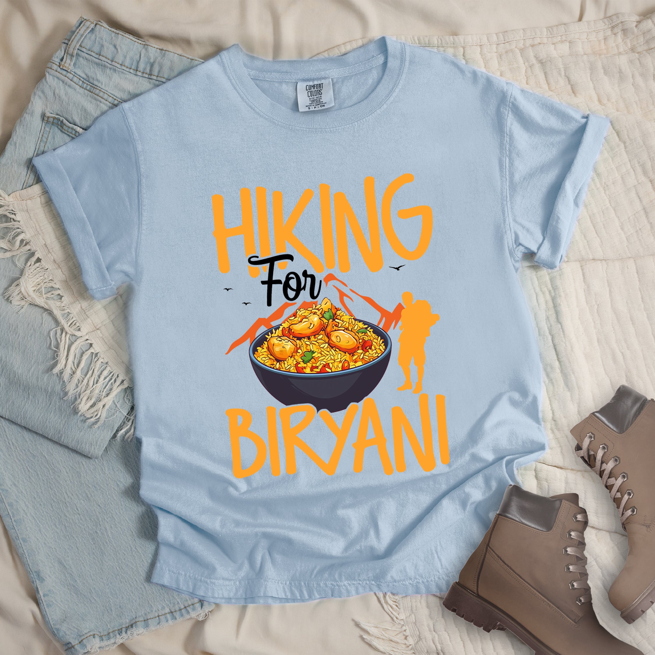 Chambray (Light Blue) t-shirt with orange text reading "HIKING For BIRYANI". "HIKING" appears in large orange letters at the top, "For" in black script in the middle, and "BIRYANI" in orange at the bottom. Features a detailed illustration of a blue bowl filled with yellow biryani rice and meat pieces in the center, bright orange mountain silhouettes in the background, and an orange hiker silhouette on the right side.