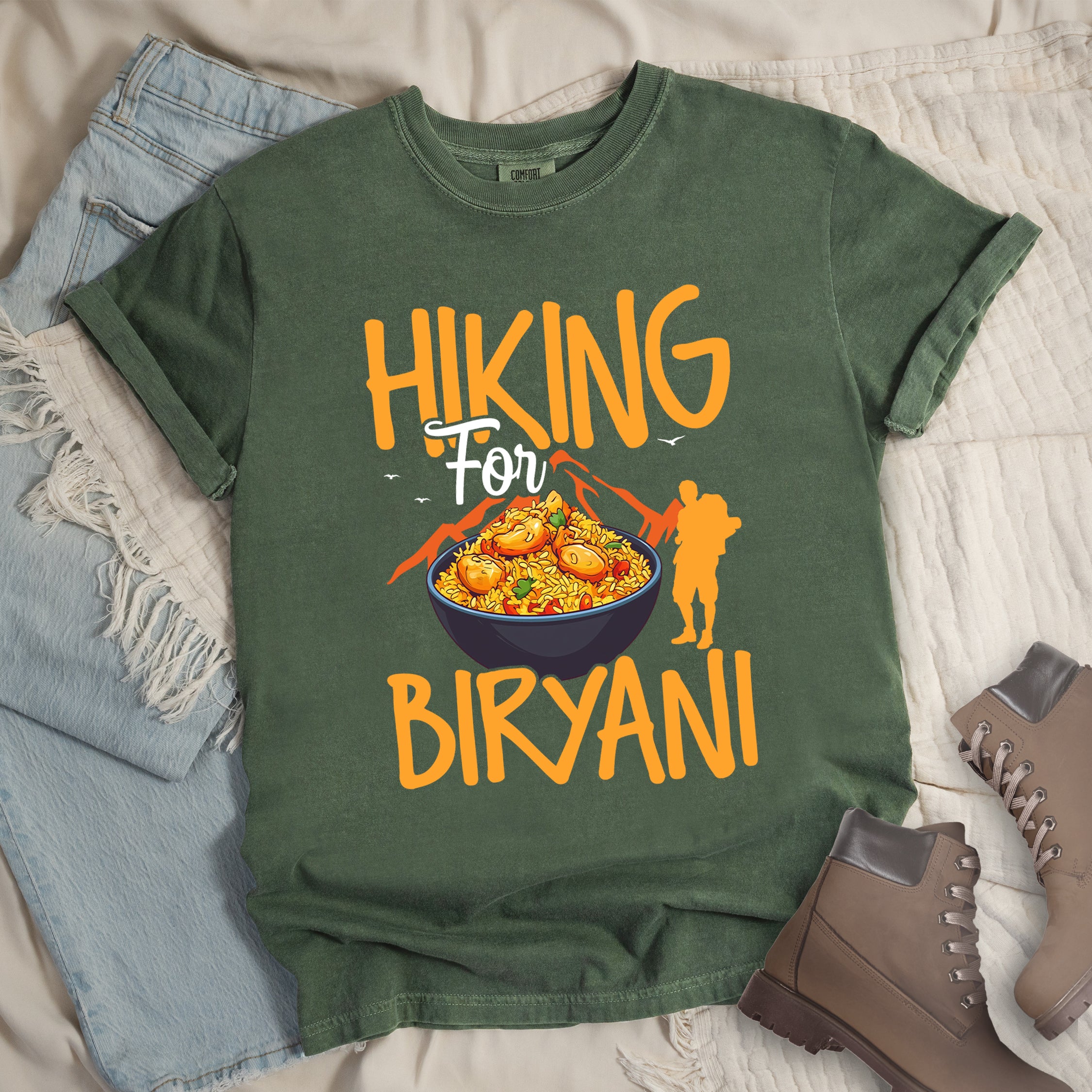 Moss (Dark Green) t-shirt with orange text reading "HIKING For BIRYANI". "HIKING" appears in large orange letters at the top, "For" in white script in the middle, and "BIRYANI" in orange at the bottom. Features a detailed illustration of a blue bowl filled with yellow biryani rice and meat pieces in the center, bright orange mountain silhouettes in the background, and an orange hiker silhouette on the right side.