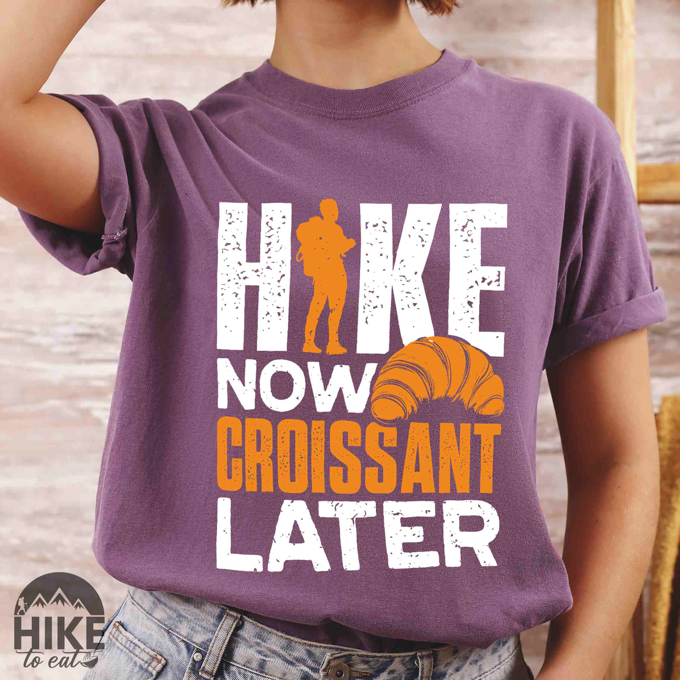 Woman wearing berry "Hike now croissant later" colored vintage tee with bold typography, birds, mountains, illustrated baked croissant, and backpacker for pastry enthusiasts.