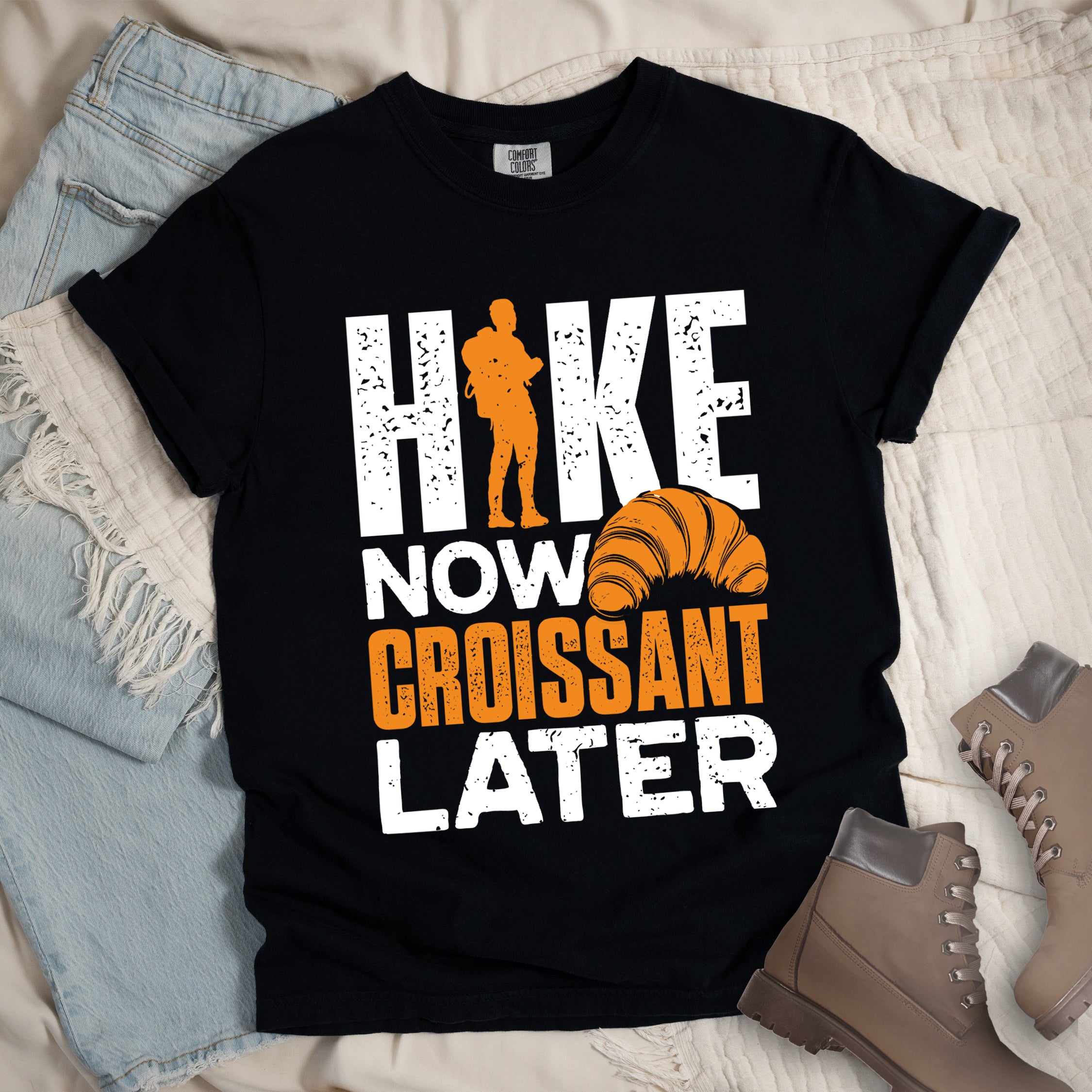 Black t-shirt with white and orange distressed text reading "HIKE NOW CROISSANT LATER". "HIKE" appears in large white letters at the top with an orange hiker silhouette replacing the letter "I", "NOW" in white in the middle, "CROISSANT" in orange, and "LATER" in white at the bottom. Features an illustration of an orange croissant between "NOW" and "LATER".  Features a classic crew neck and casual rolled sleeves, styled alongside light wash denim and brown hiking boots. 