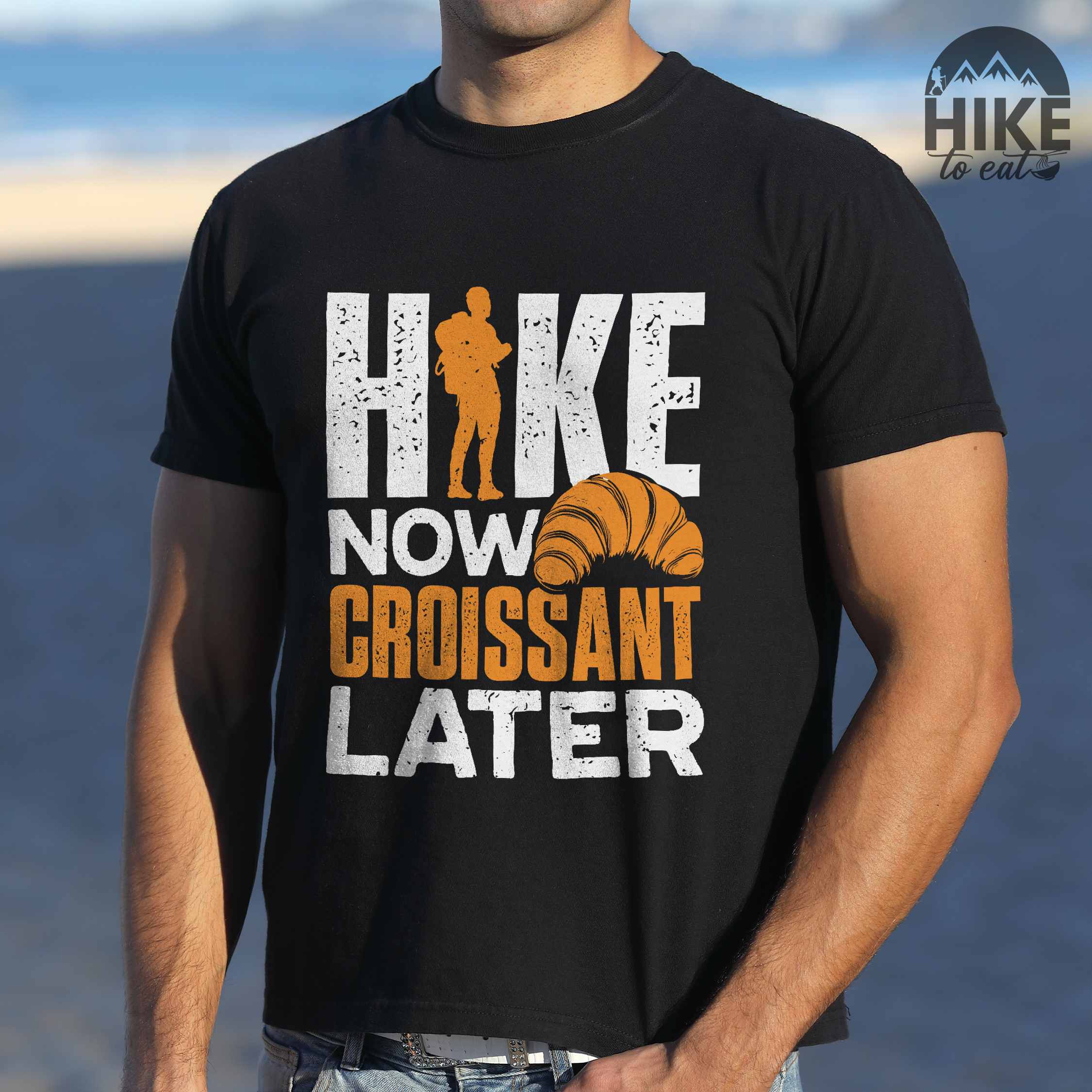 Man wearing black "Hike now croissant later" colored vintage tee with bold typography, birds, mountains, illustrated baked croissant, and backpacker for pastry enthusiasts.