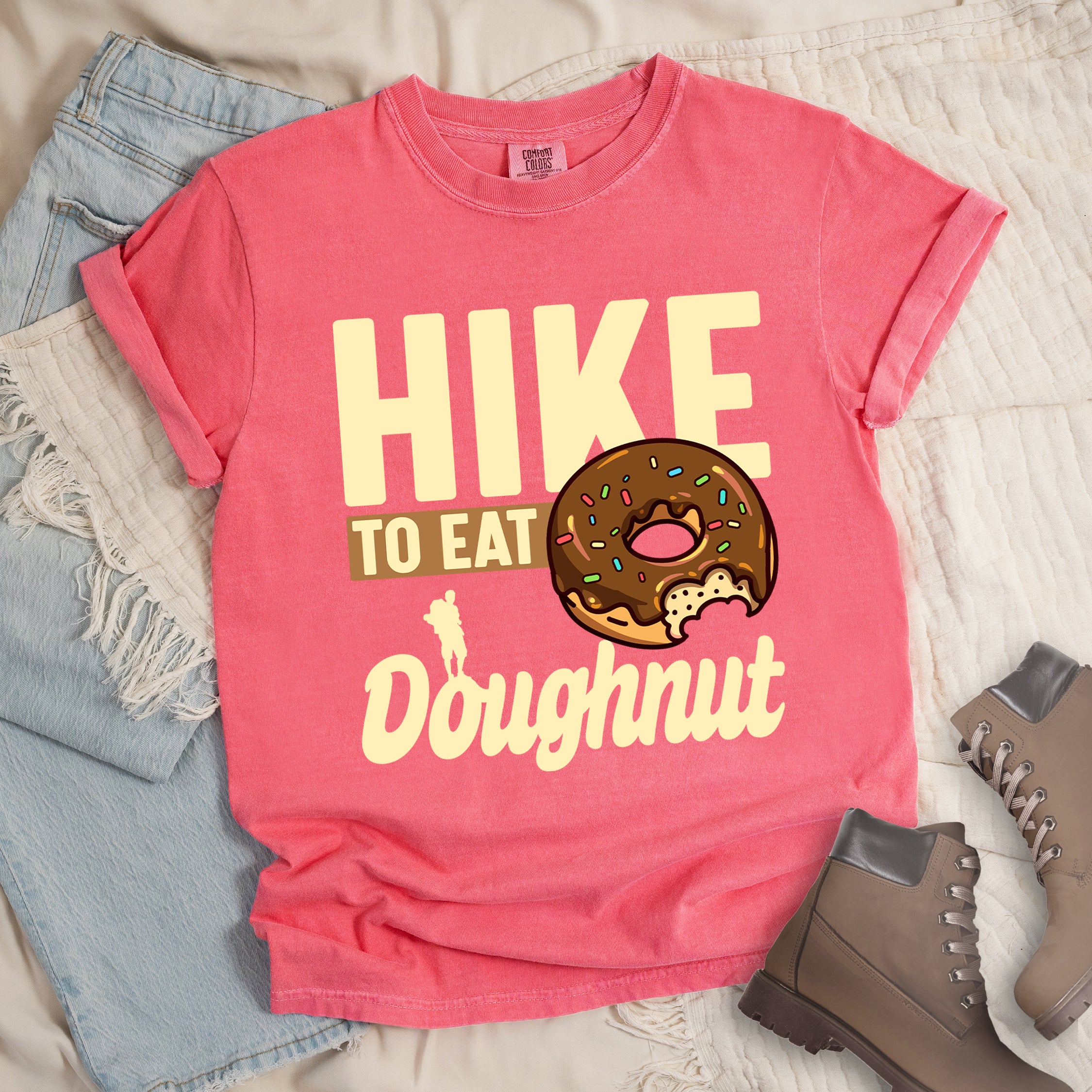 Watermelon (Light Coral) t-shirt with cream and tan text reading "HIKE TO EAT Doughnut". "HIKE" appears in large cream letters at the top, "TO EAT" on a tan rectangular background in the middle, and "Doughnut" in cream script at the bottom. Features a chocolate frosted doughnut with colorful sprinkles and a bite taken out of it on the right side, and a small cream silhouette of a hiker on the left. 