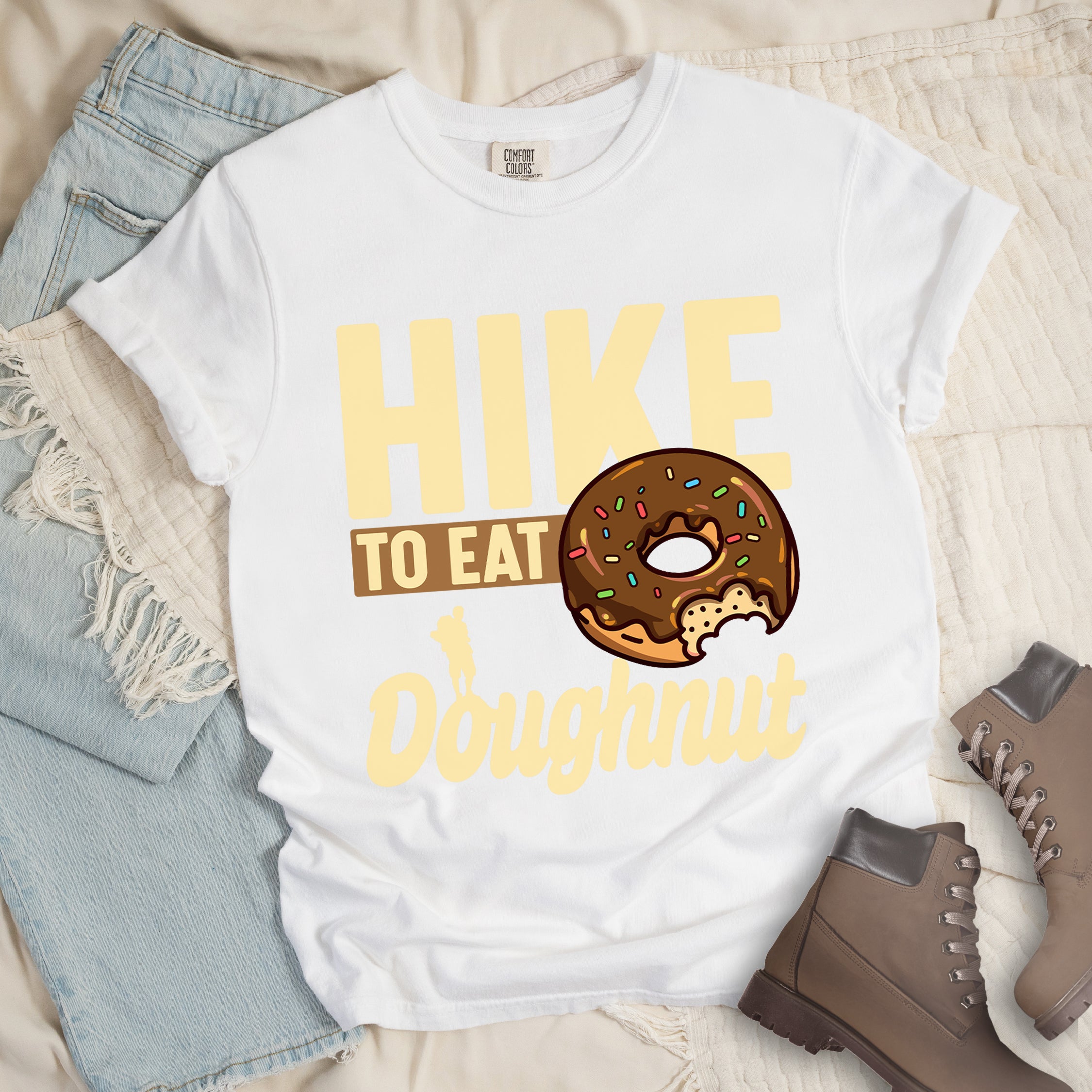 White t-shirt with cream and tan text reading "HIKE TO EAT Doughnut". "HIKE" appears in large cream letters at the top, "TO EAT" on a tan rectangular background in the middle, and "Doughnut" in cream script at the bottom. Features a chocolate frosted doughnut with colorful sprinkles and a bite taken out of it on the right side, and a small cream silhouette of a hiker on the left. 
