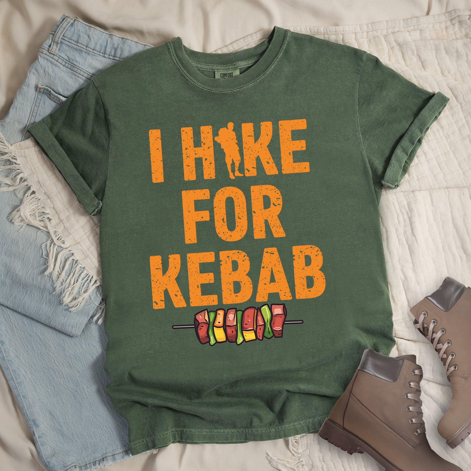 Moss (Dark Green) t-shirt with orange distressed text reading "I HIKE FOR KEBAB". "I HIKE" appears at the top with a hiker silhouette integrated into the letter "I", "FOR" in the middle, and "KEBAB" at the bottom. Below the text is a colorful illustration of a kebab skewer with meat and vegetables. Features a classic crew neck and casual rolled sleeves, styled alongside light wash denim and brown hiking boots. 