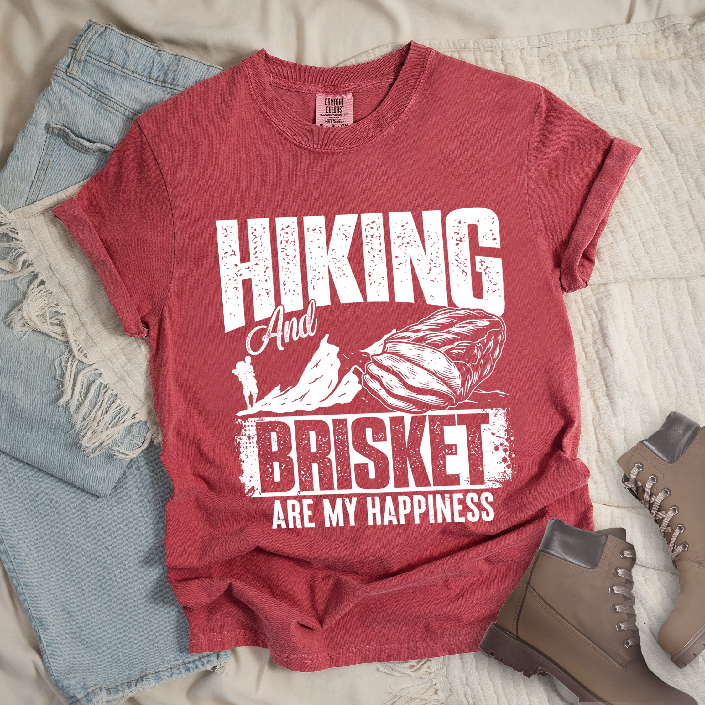 Crimson (Dark Pink) t-shirt with white distressed text reading "HIKING And BRISKET ARE MY HAPPINESS". "HIKING" appears in large letters at the top, "And" in script in the middle, "BRISKET" in bold distressed letters, and "ARE MY HAPPINESS" in smaller text at the bottom. Features a detailed white illustration of a brisket cut of meat on the right and mountain silhouettes with a small hiker figure on the left.