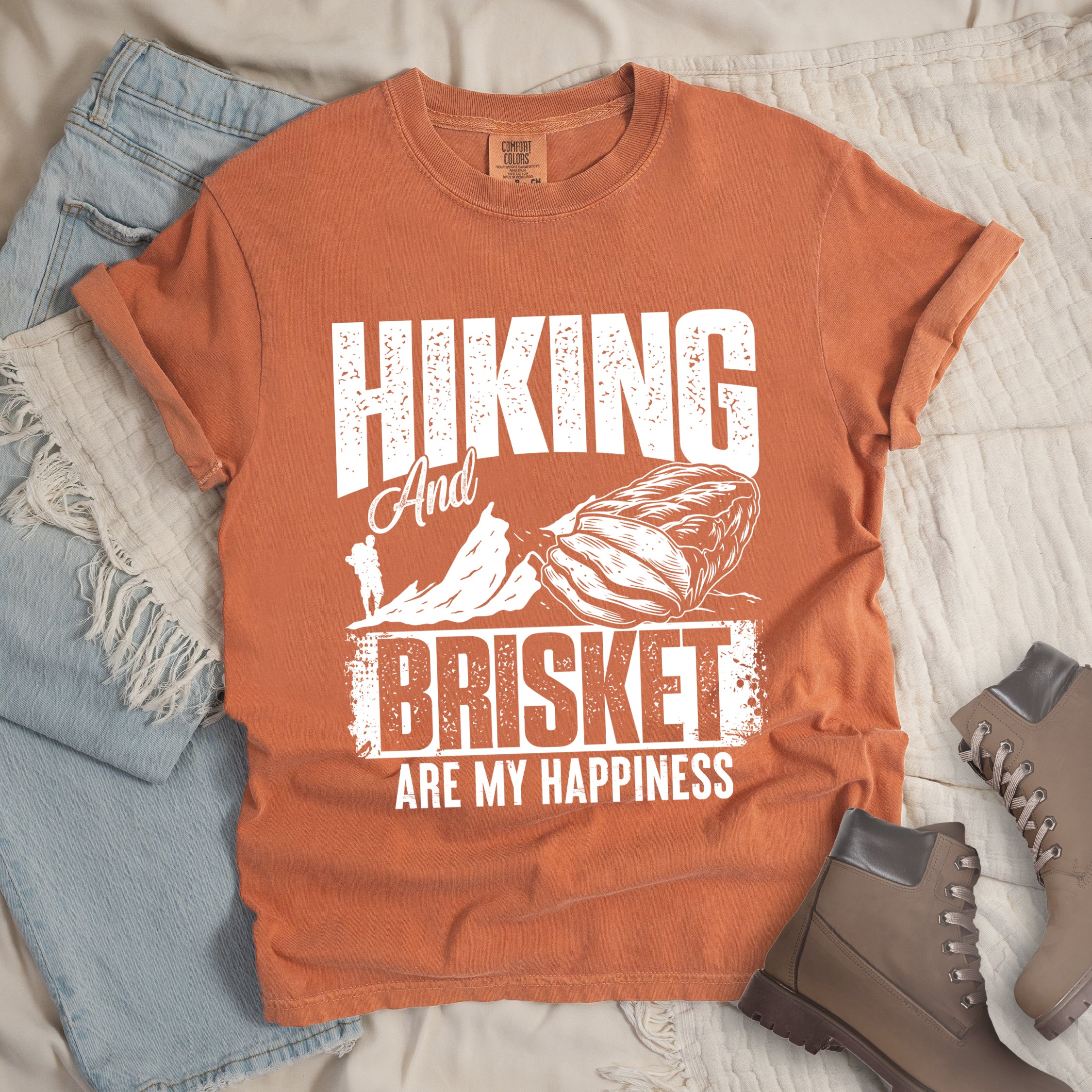 Yam (Brown) t-shirt with white distressed text reading "HIKING And BRISKET ARE MY HAPPINESS". "HIKING" appears in large letters at the top, "And" in script in the middle, "BRISKET" in bold distressed letters, and "ARE MY HAPPINESS" in smaller text at the bottom. Features a detailed white illustration of a brisket cut of meat on the right and mountain silhouettes with a small hiker figure on the left. 