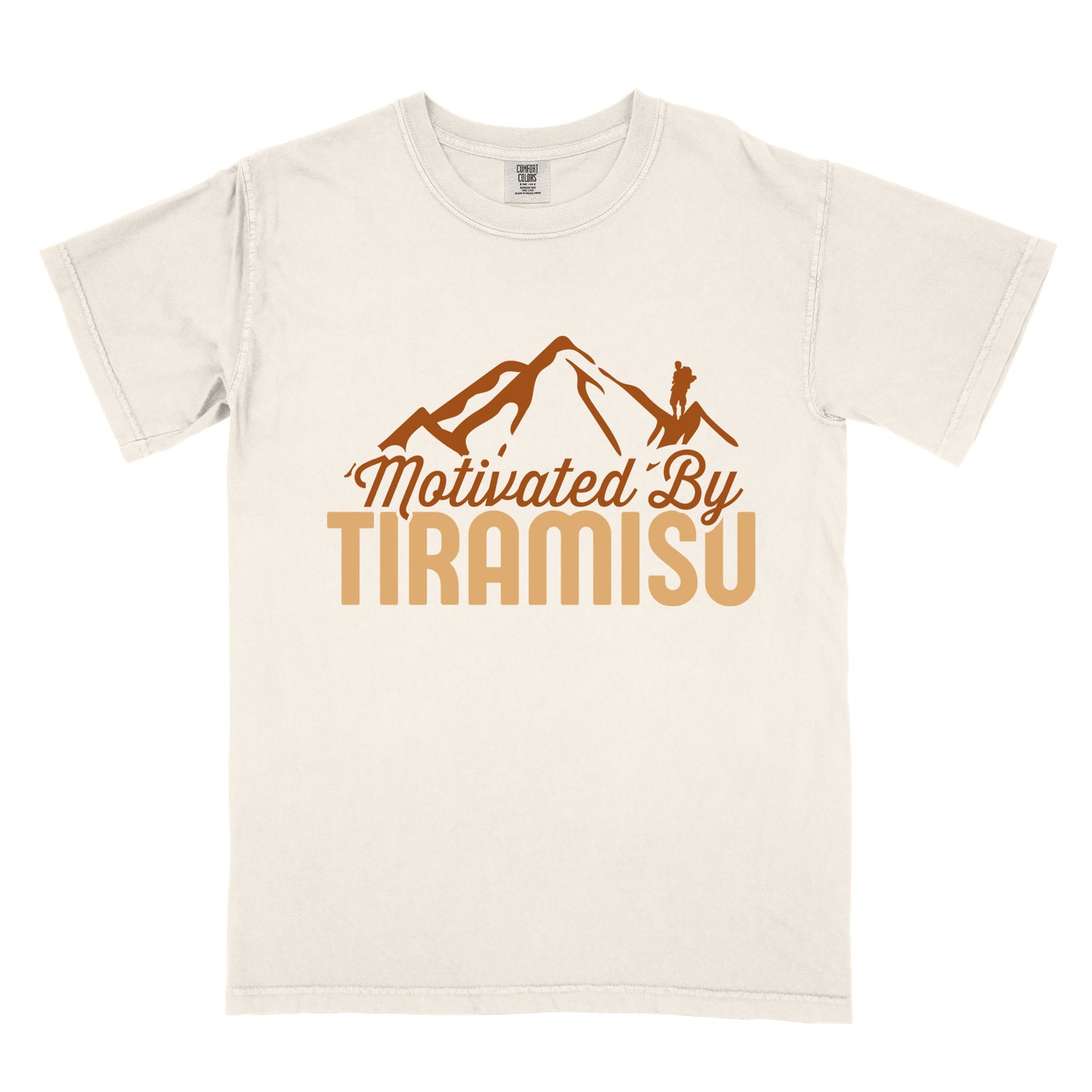 Ivory (Cream White) t-shirt with brown and gold text reading "Motivated By TIRAMISU". "Motivated By" appears in brown script underneath a brown mountain silhouette with a small hiker figure on the peak, and "TIRAMISU" in large gold letters at the bottom. The design humorously connects the Italian coffee-flavored dessert as motivation for hiking and outdoor activities.