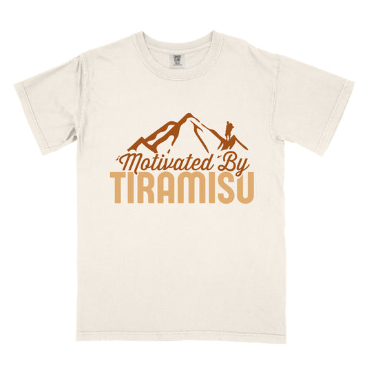 Ivory (Cream White) t-shirt with brown and gold text reading "Motivated By TIRAMISU". "Motivated By" appears in brown script underneath a brown mountain silhouette with a small hiker figure on the peak, and "TIRAMISU" in large gold letters at the bottom. The design humorously connects the Italian coffee-flavored dessert as motivation for hiking and outdoor activities.