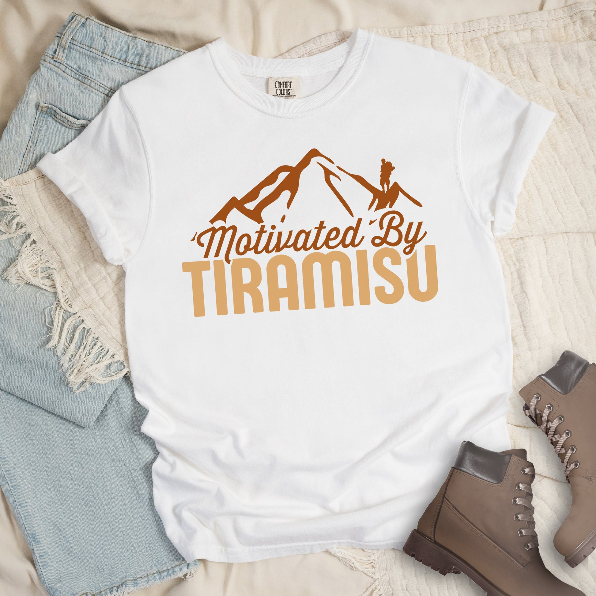 White t-shirt with brown and gold text reading "Motivated By TIRAMISU". "Motivated By" appears in brown script underneath a brown mountain silhouette with a small hiker figure on the peak, and "TIRAMISU" in large gold letters at the bottom. The shirt features a classic crew neck and casual rolled sleeves, styled alongside light wash denim and brown hiking boots. The design humorously connects the Italian coffee-flavored dessert as motivation for hiking and outdoor activities. 