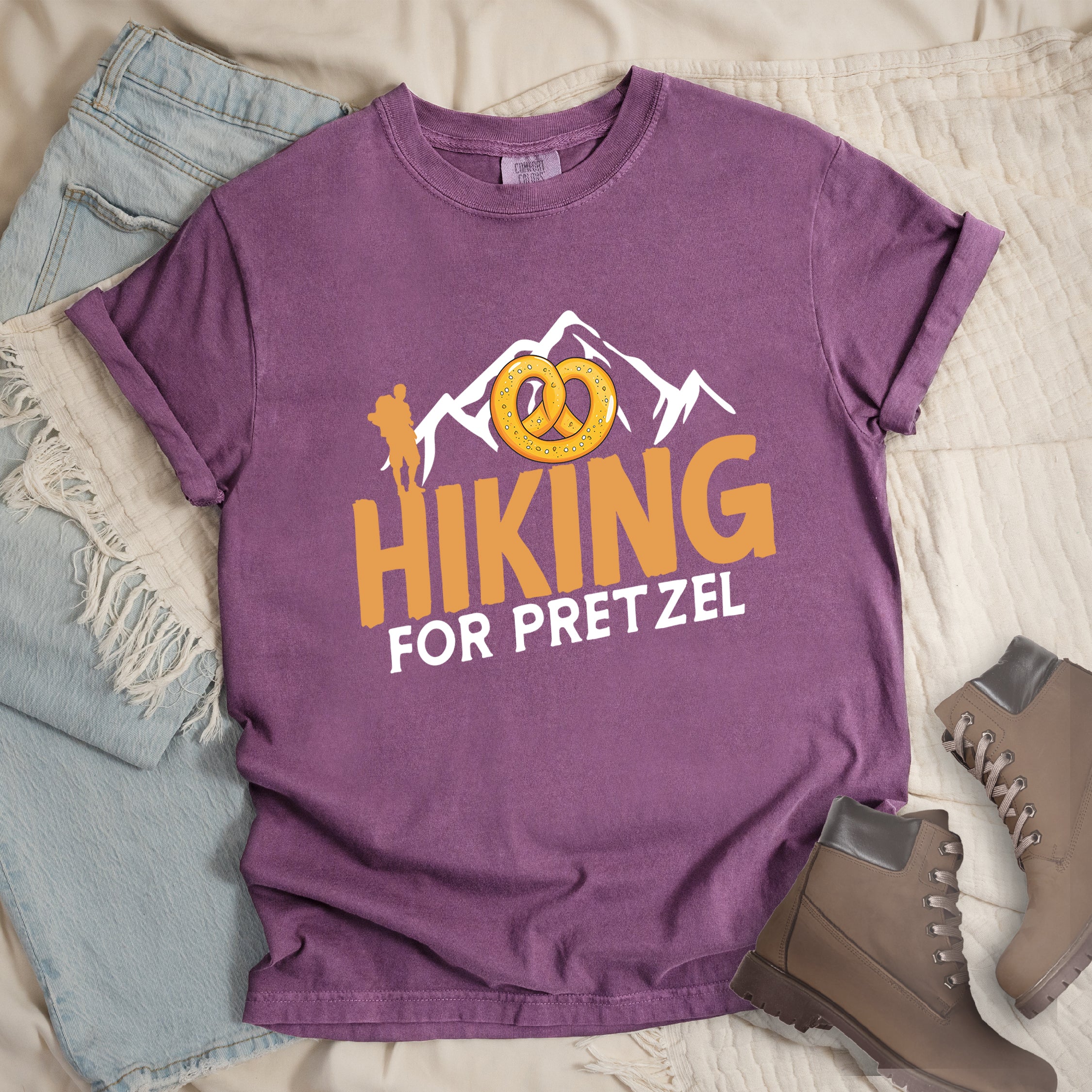 Berry (Dark Violet) t-shirt with orange and white text reading "HIKING FOR PRETZEL". "HIKING" appears in large orange letters in the middle, and "FOR PRETZEL" in white below it. Features white mountain silhouettes in the background, an orange hiker silhouette on the left side, and an illustration of a golden pretzel with salt above the word "HIKING". The design connects hiking activity with pretzel snacks as a reward.