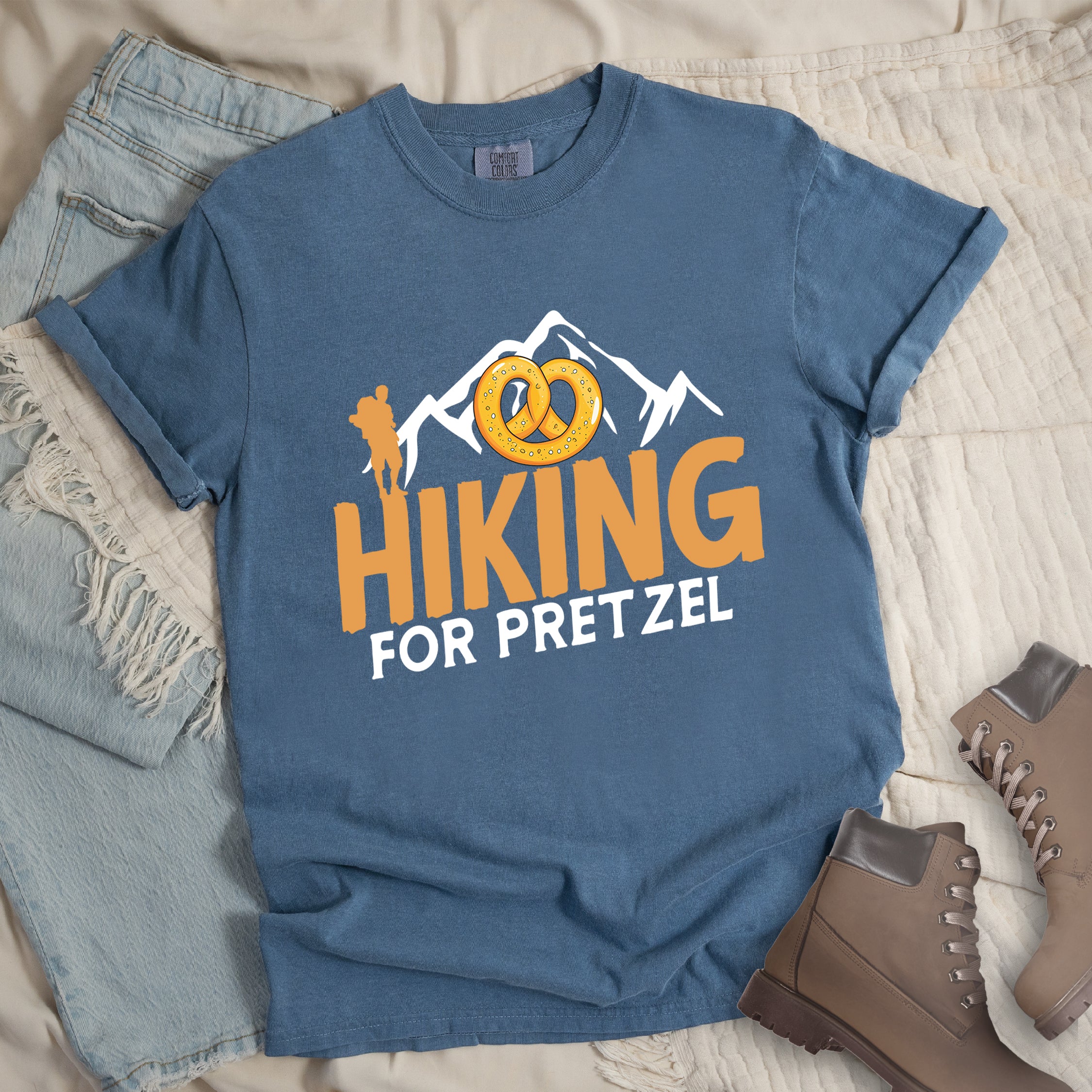 Blue Jean (Denim Blue) t-shirt with orange and white text reading "HIKING FOR PRETZEL". "HIKING" appears in large orange letters in the middle, and "FOR PRETZEL" in white below it. Features white mountain silhouettes in the background, an orange hiker silhouette on the left side, and an illustration of a golden pretzel with salt above the word "HIKING". The design connects hiking activity with pretzel snacks as a reward.