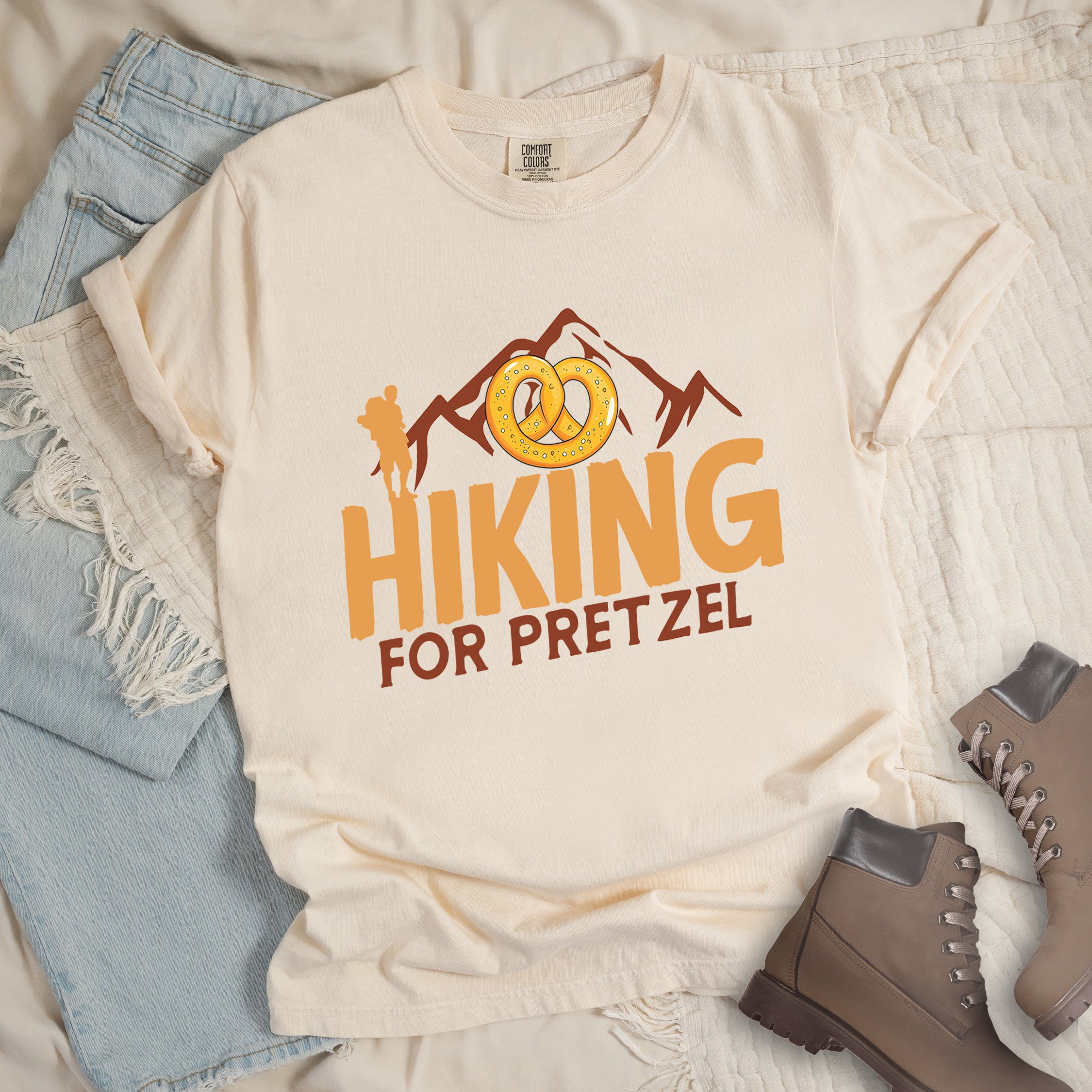 Ivory (Cream White) t-shirt with orange and brown text reading "HIKING FOR PRETZEL". "HIKING" appears in large orange letters in the middle, and "FOR PRETZEL" in brown below it. Features brown mountain silhouettes in the background, an orange hiker silhouette on the left side, and an illustration of a golden pretzel with salt above the word "HIKING". The design connects hiking activity with pretzel snacks as a reward.