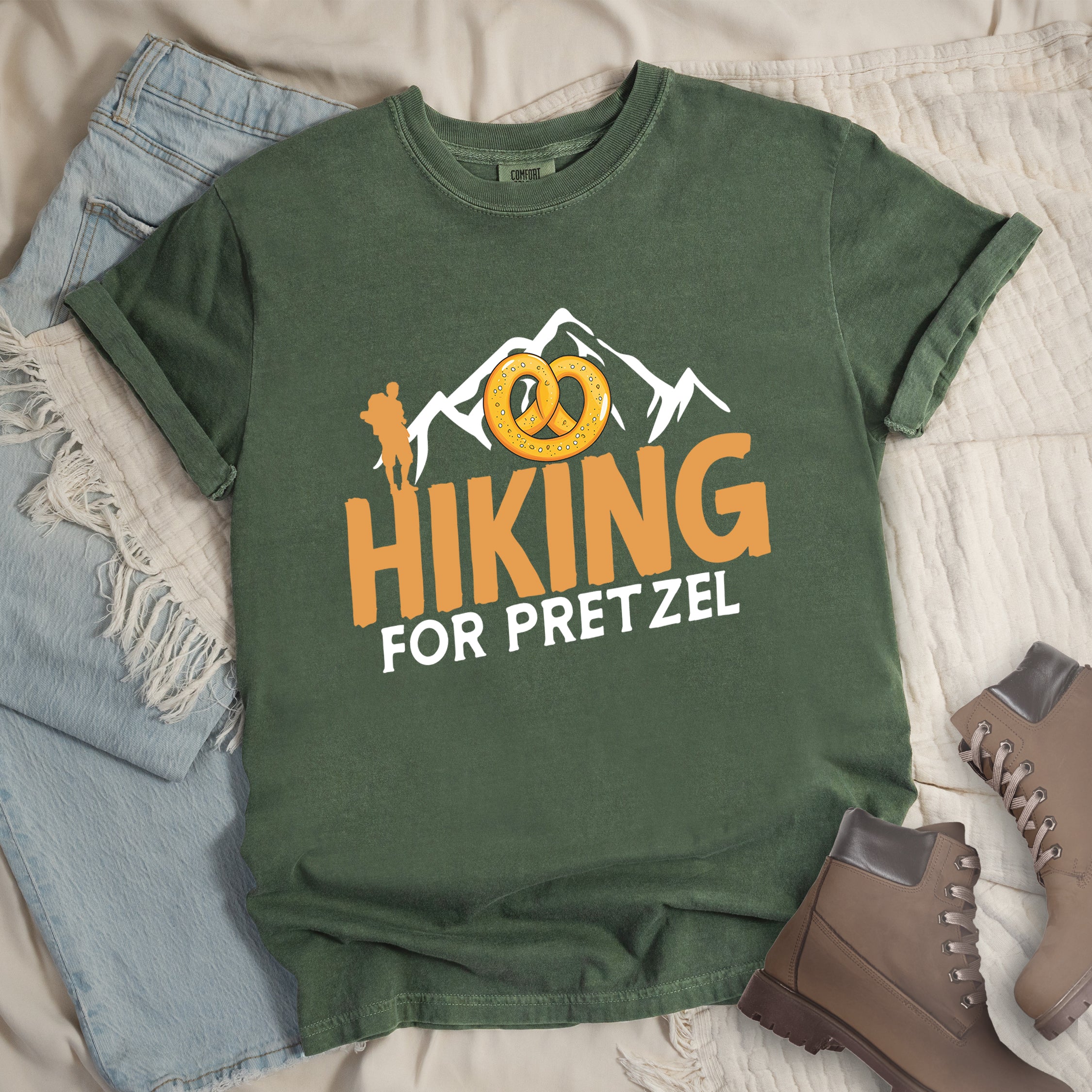 Moss (Dark Green) t-shirt with orange and white text reading "HIKING FOR PRETZEL". "HIKING" appears in large orange letters in the middle, and "FOR PRETZEL" in white below it. Features white mountain silhouettes in the background, an orange hiker silhouette on the left side, and an illustration of a golden pretzel with salt above the word "HIKING". The design connects hiking activity with pretzel snacks as a reward.