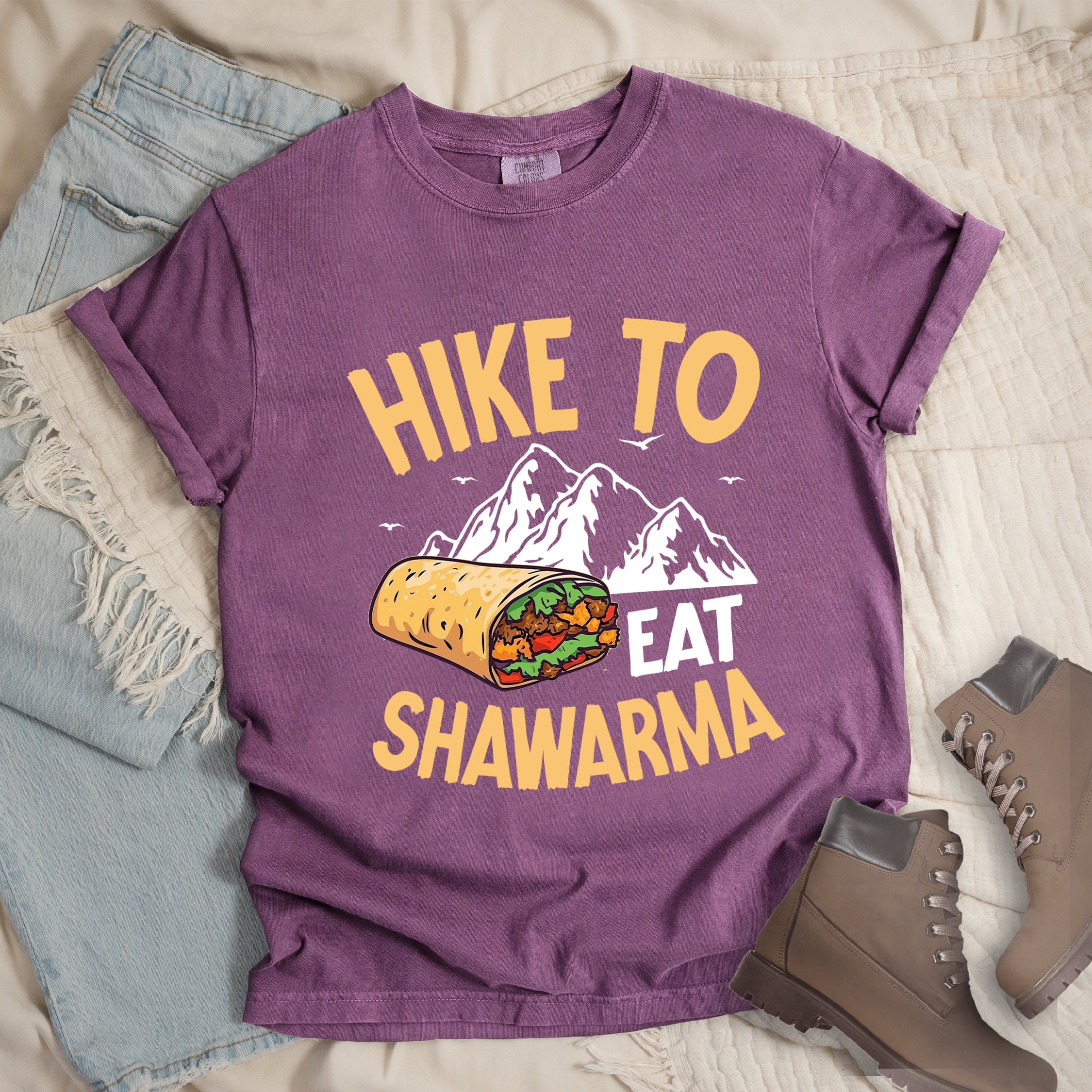 Berry (Dark Violet) t-shirt with golden yellow and white text reading "HIKE TO EAT SHAWARMA". "HIKE TO" appears in golden yellow letters at the top, "EAT" in white in the middle, and "SHAWARMA" in golden yellow at the bottom. Features a detailed illustration of a shawarma wrap with visible meat and vegetables, white mountain silhouettes with small bird silhouettes in the background. Features a classic crew neck and casual rolled sleeves, styled alongside light wash denim and brown hiking boots. 