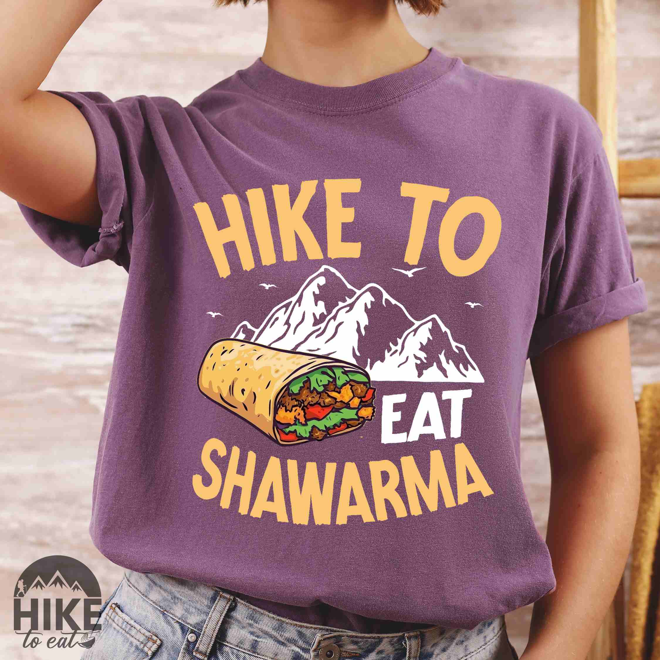 Woman wearing berry "Hike to Eat Shawarma" colored vintage tee with bold typography, mountains, birds, and illustrated shawarma, and backpackers for shawarma lovers.
