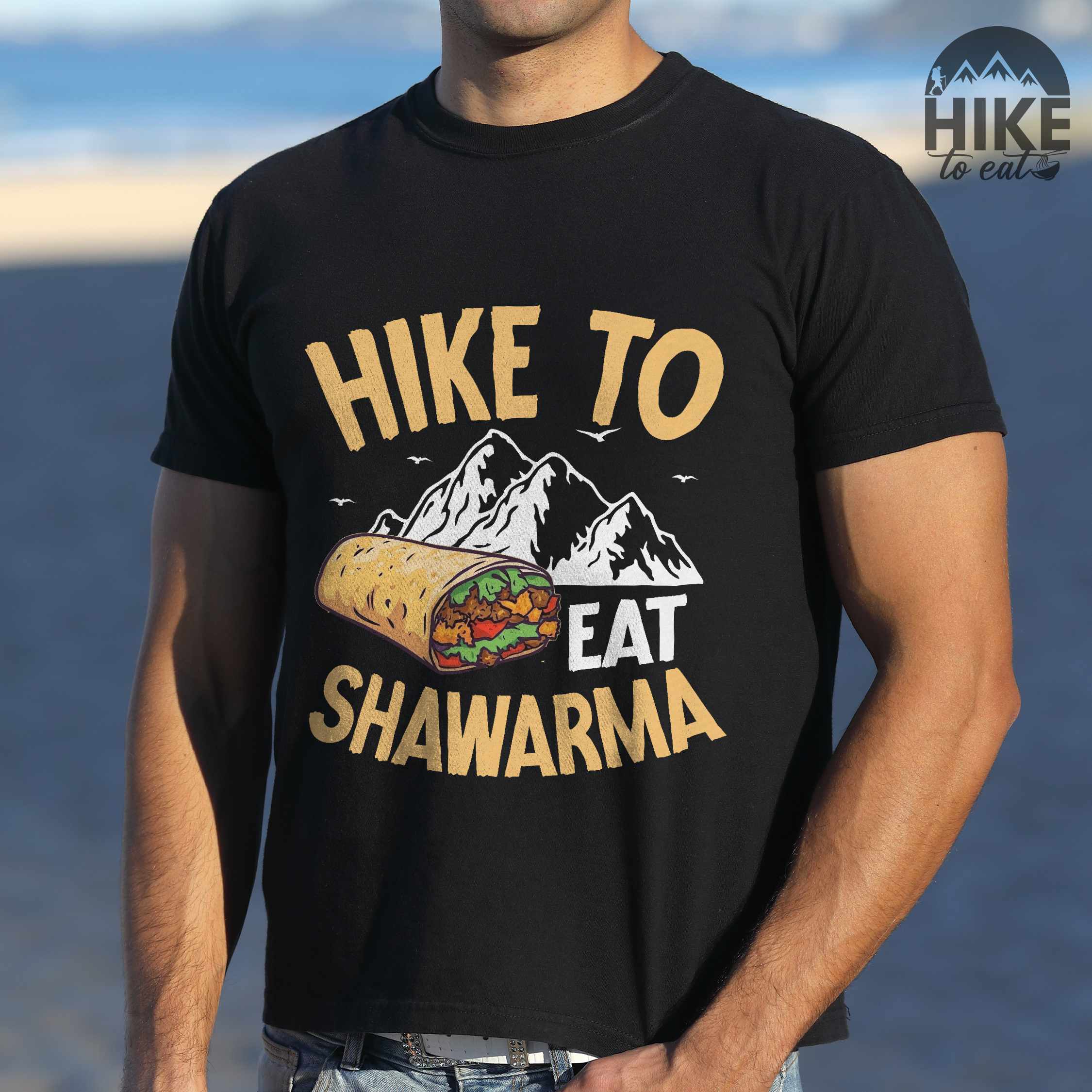 Man wearing black "Hike to Eat Shawarma" colored vintage tee with bold typography, mountains, birds, and illustrated shawarma, and backpackers for shawarma lovers.