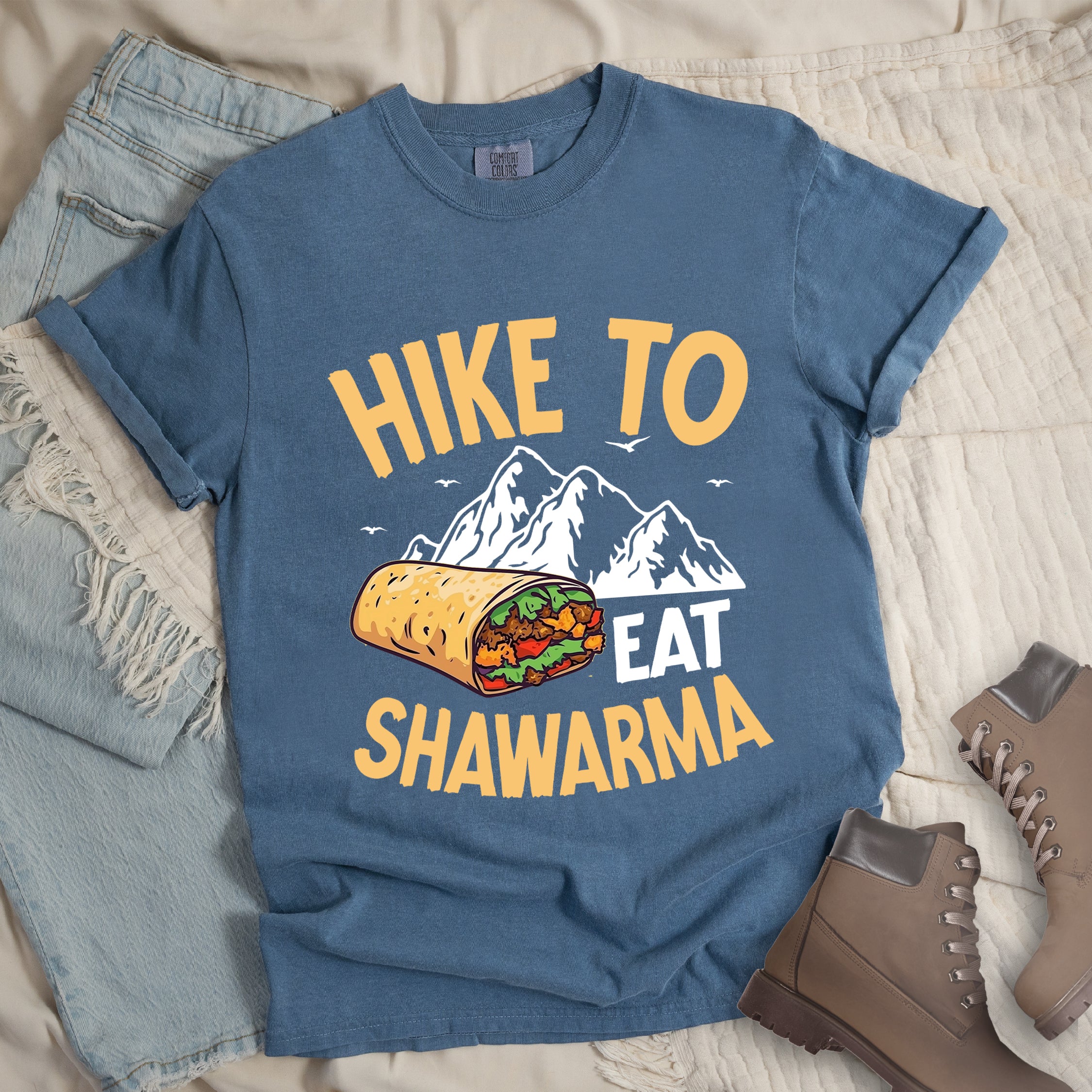 Blue Jean (Denim Blue) t-shirt with golden yellow and white text reading "HIKE TO EAT SHAWARMA". "HIKE TO" appears in golden yellow letters at the top, "EAT" in white in the middle, and "SHAWARMA" in golden yellow at the bottom. Features a detailed illustration of a shawarma wrap with visible meat and vegetables, white mountain silhouettes with small bird silhouettes in the background. Features a classic crew neck and casual rolled sleeves, styled alongside light wash denim and brown hiking boots. 