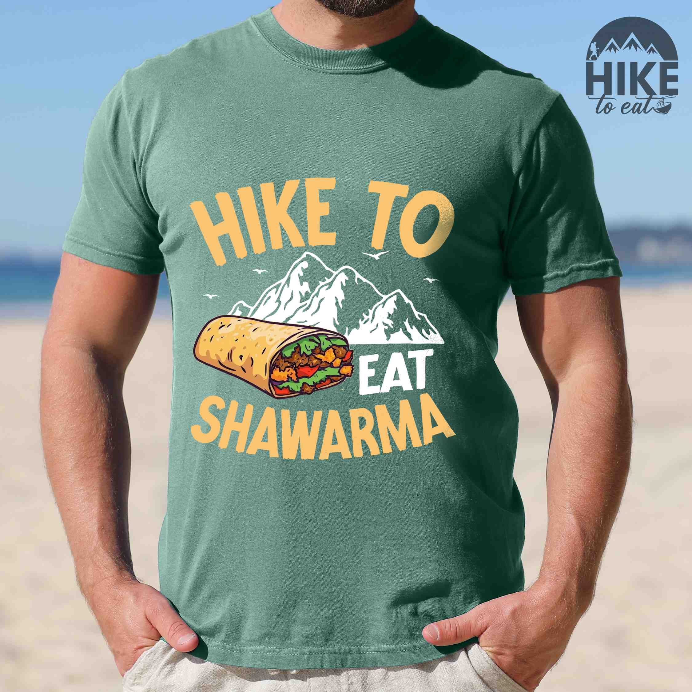 Man wearing light green "Hike to Eat Shawarma" colored vintage tee with bold typography, mountains, birds, and illustrated shawarma, and backpackers for shawarma lovers.