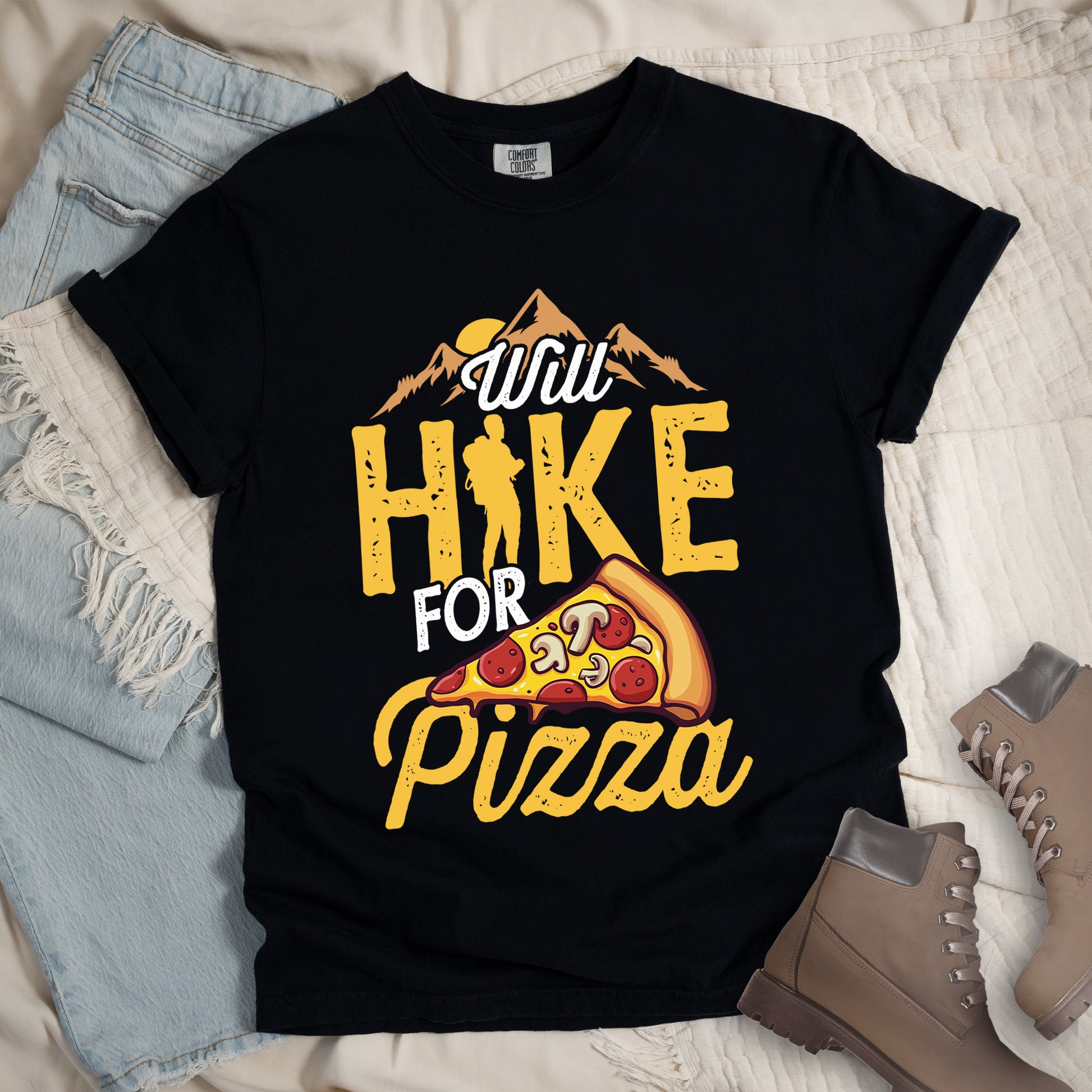 Black t-shirt with "Will HIKE FOR pizza" design. Features "Will" in white script above mountain graphics with a yellow sun. "HIKE" appears in large yellow distressed letters with a hiker silhouette replacing the letter "I". "FOR" in white text sits above "pizza" in yellow script. An illustrated pizza slice with pepperoni and mushrooms appears between "FOR" and "pizza". The shirt features a classic crew neck and casual rolled sleeves, styled alongside light wash denim and brown hiking boots.