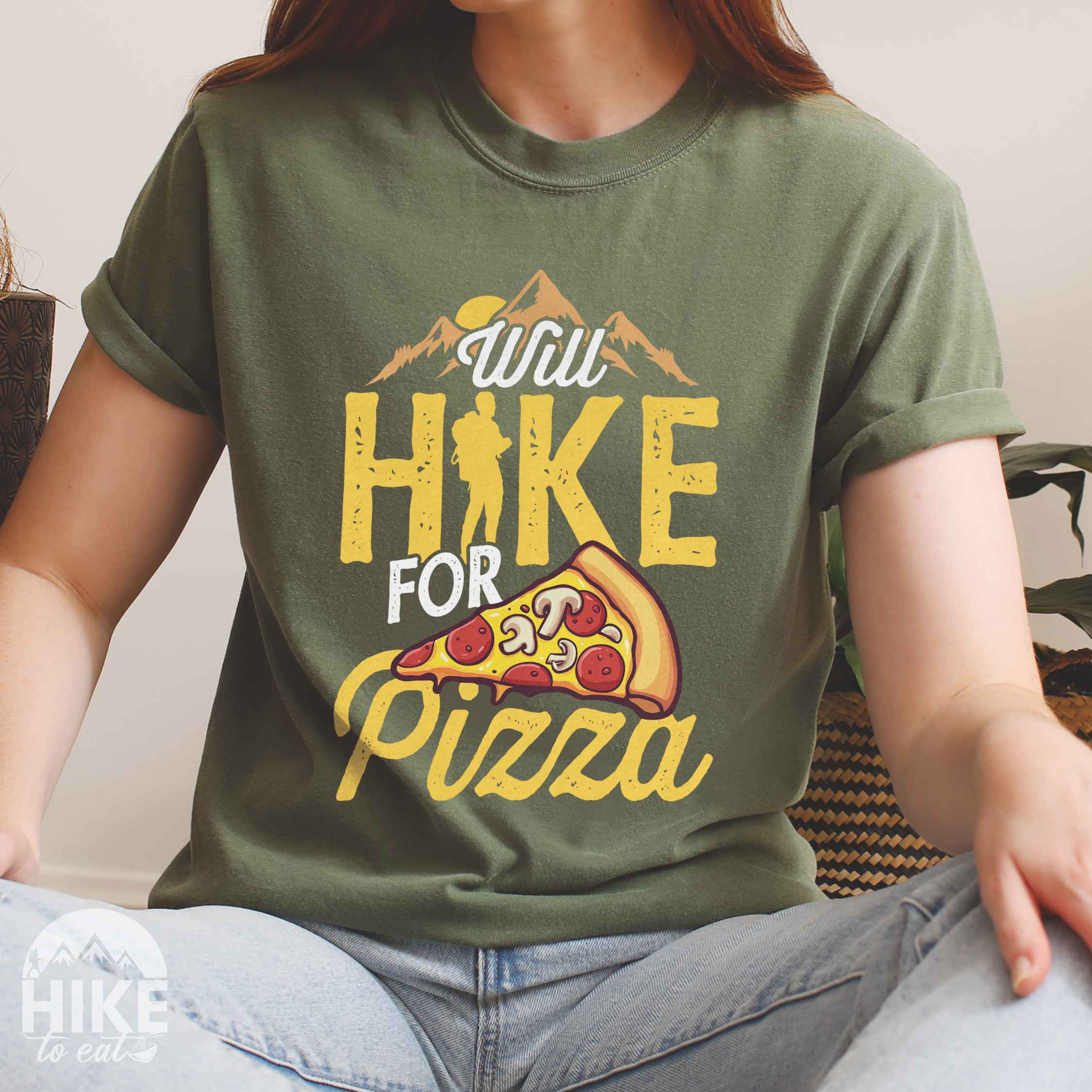 Woman wearing moss colored vintage tee featuring Hiking design of mountains with a sunrise behind and a slice of pizza in front of the phrase 'Will Hike for Pizza'.