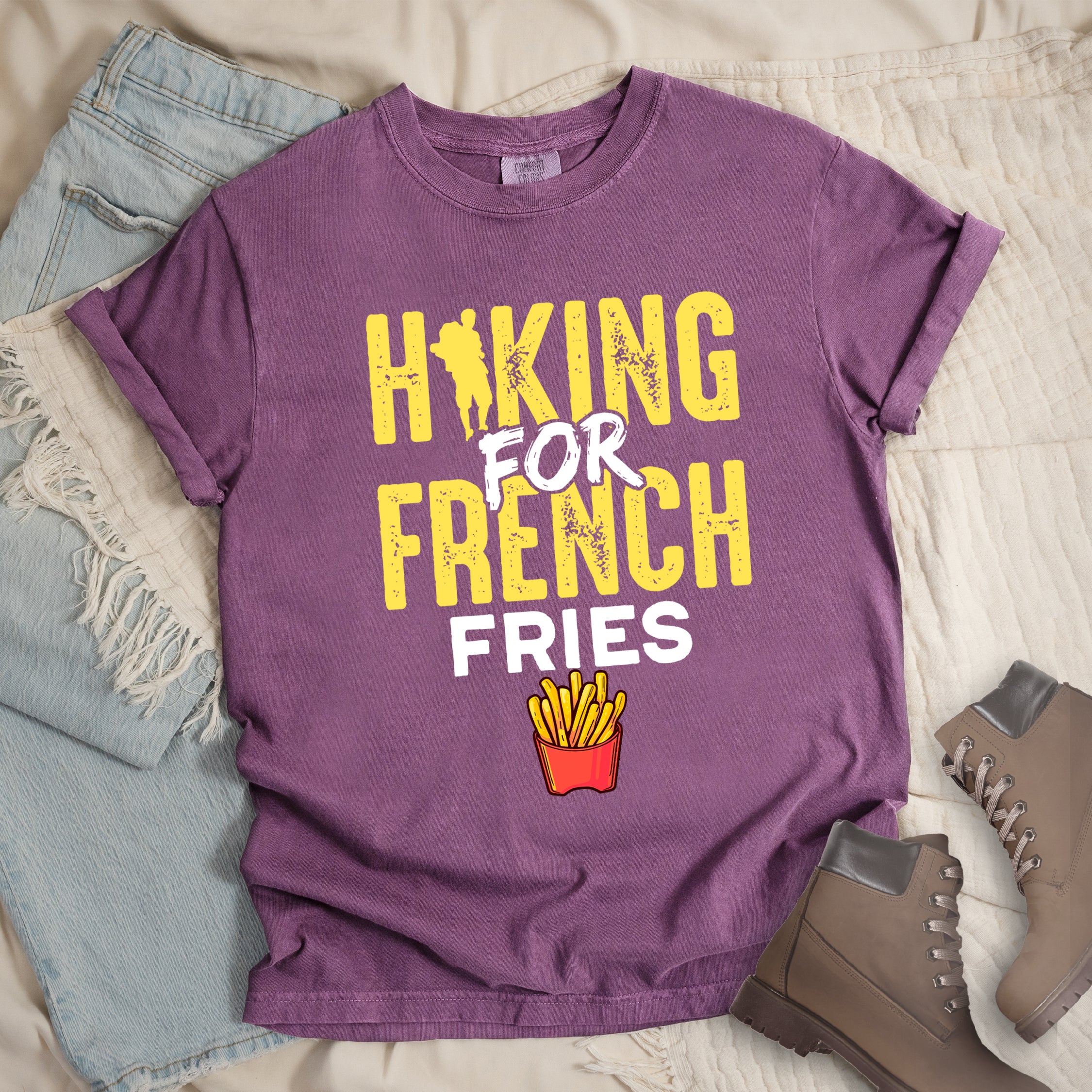 Berry (Dark Violet) t-shirt with yellow and white distressed text reading "HIKING FOR FRENCH FRIES". "HIKING" appears in yellow at the top with a hiker silhouette integrated into the letter "I", "FOR" in white brushstroke text in the middle, "FRENCH" in yellow, and "FRIES" in white at the bottom. Features an illustration of french fries in a red container below the text. Features a classic crew neck and casual rolled sleeves, styled alongside light wash denim and brown hiking boots. 