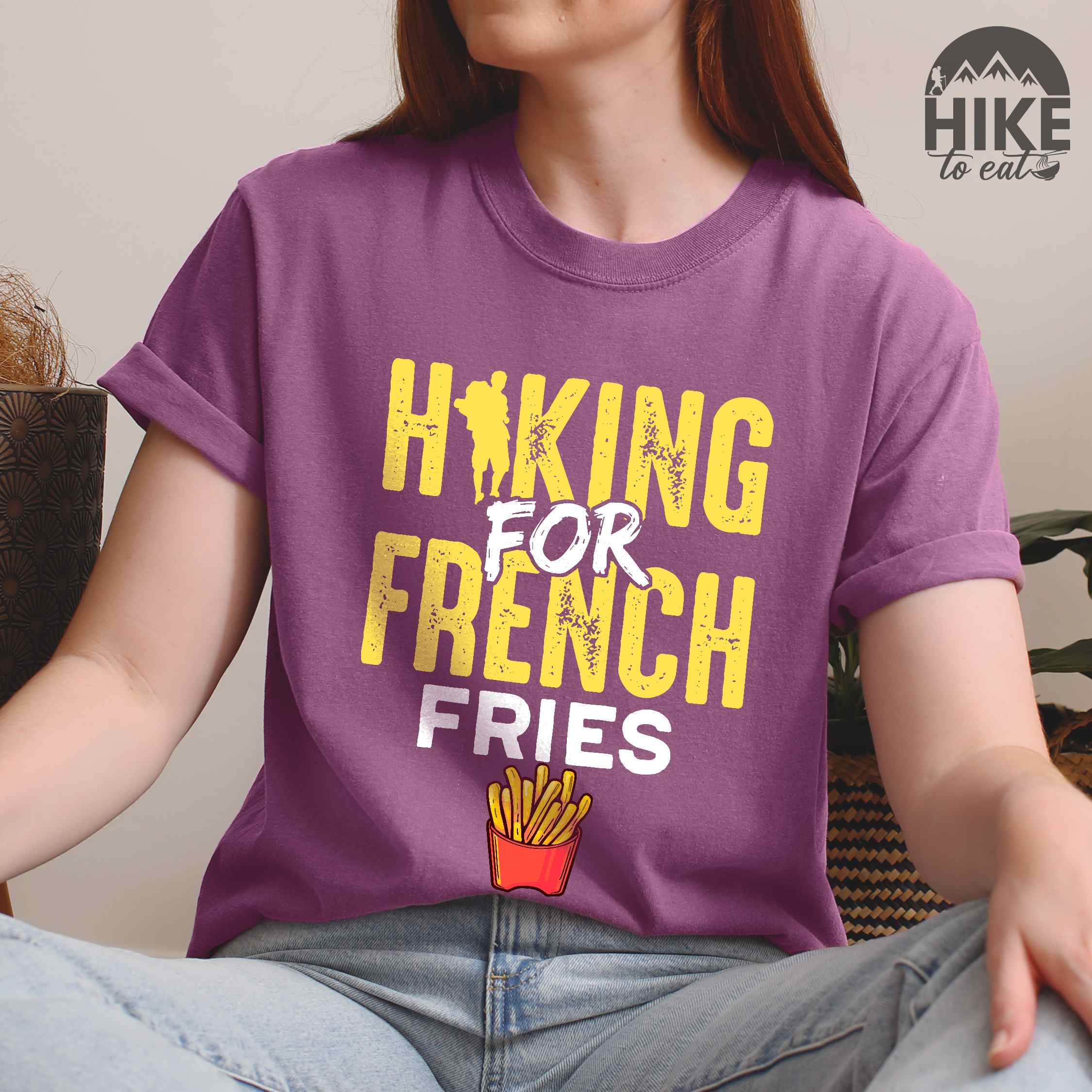 woman wearing berry "Hiking for french fries" colored vintage tee with bold typography, mountains, illustrated fries, and backpacker for comfort food enthusiasts.