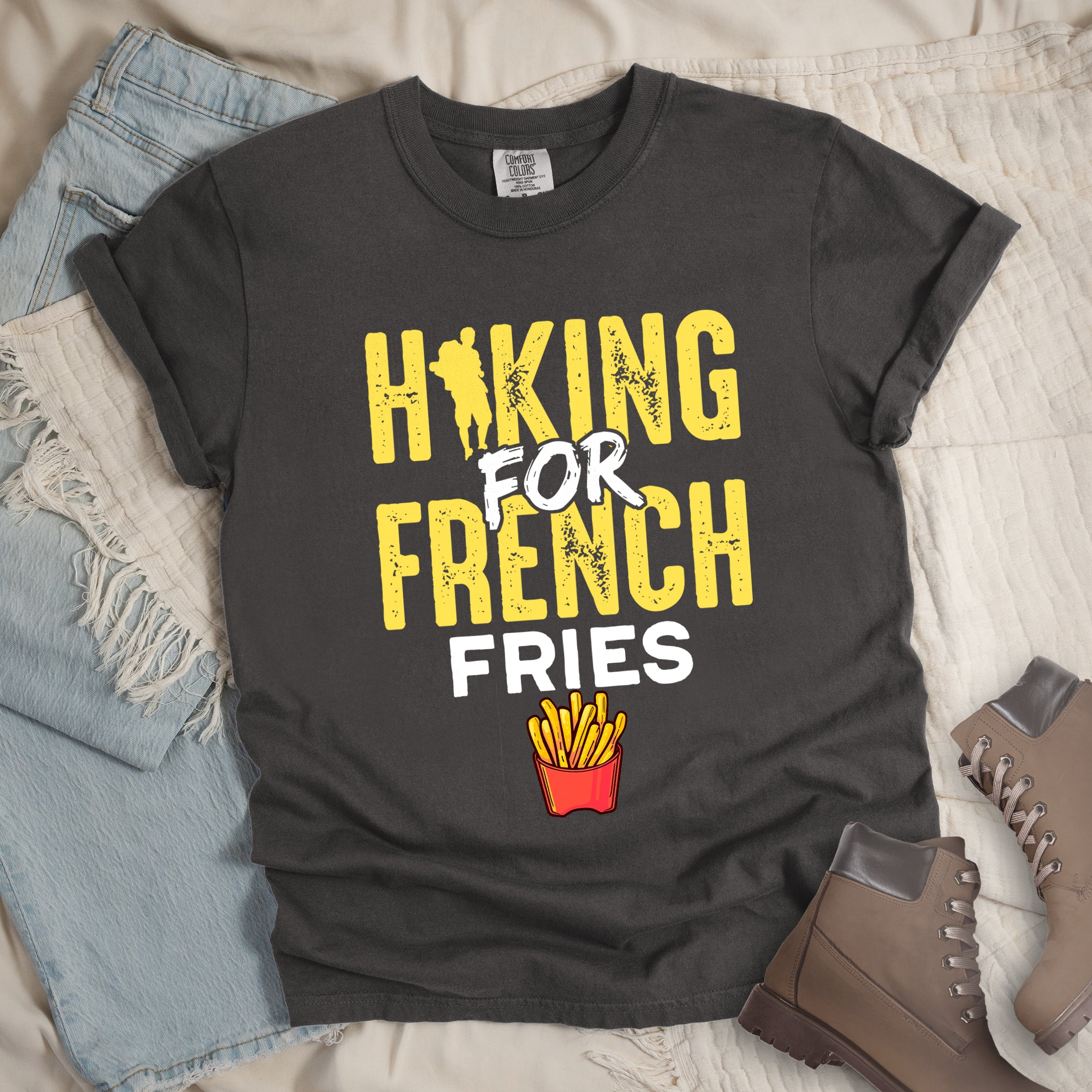 Pepper (Dark Gray) t-shirt with yellow and white distressed text reading "HIKING FOR FRENCH FRIES". "HIKING" appears in yellow at the top with a hiker silhouette integrated into the letter "I", "FOR" in white brushstroke text in the middle, "FRENCH" in yellow, and "FRIES" in white at the bottom. Features an illustration of french fries in a red container below the text. Features a classic crew neck and casual rolled sleeves, styled alongside light wash denim and brown hiking boots. 