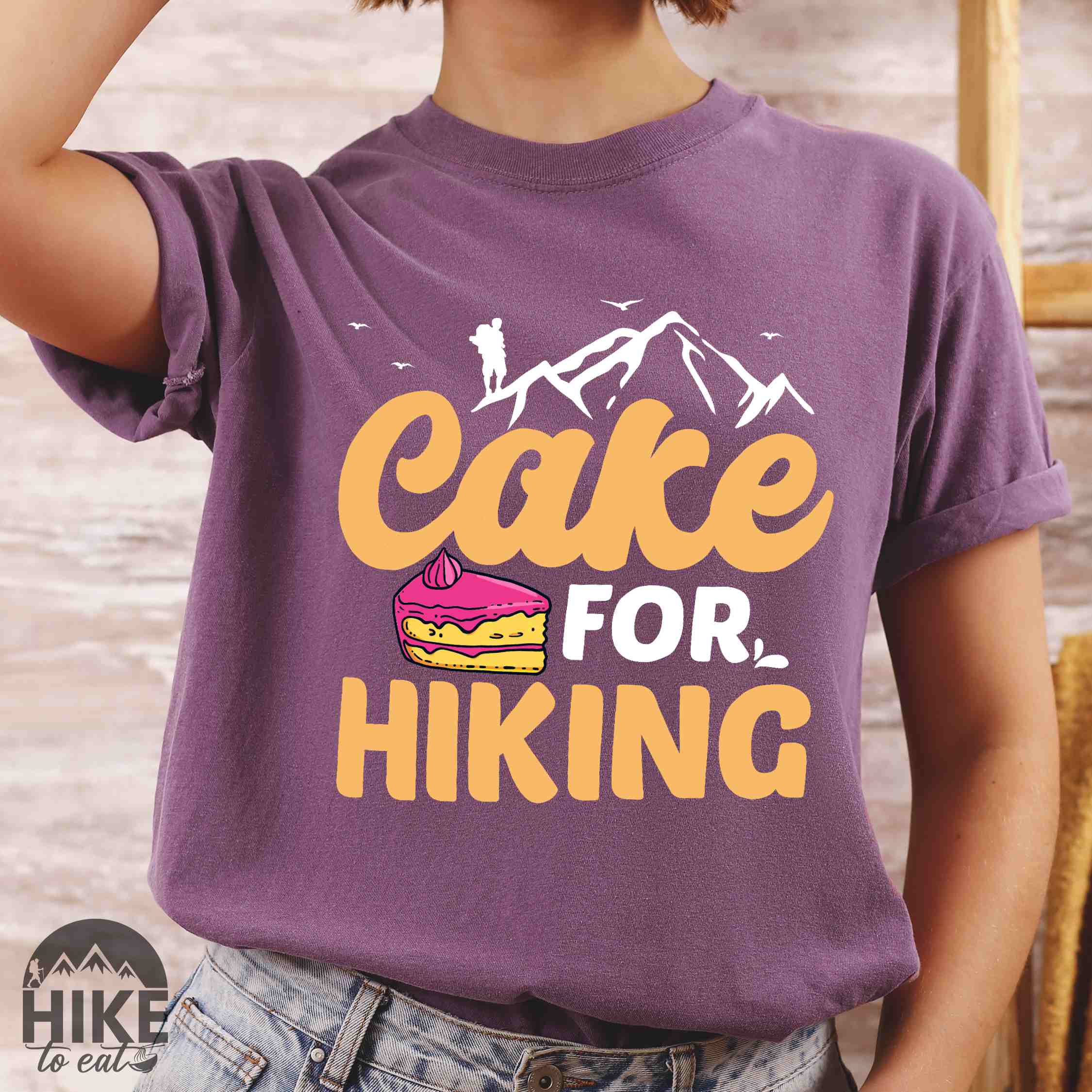 Woman wearing berry "Cake for Hiking" colored vintage tee with bold typography, mountains, illustrated a slice of cake, and backpackers with a sweet tooth.