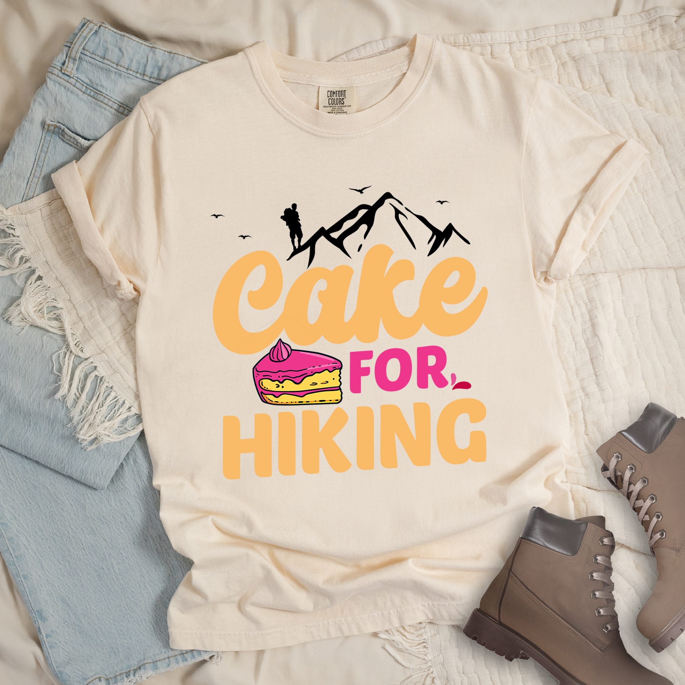 Ivory t-shirt with text reading "Cake FOR HIKING". Features white mountain silhouettes with a hiker figure and birds at the top, and an illustration of a yellow cake slice with pink frosting between the words.