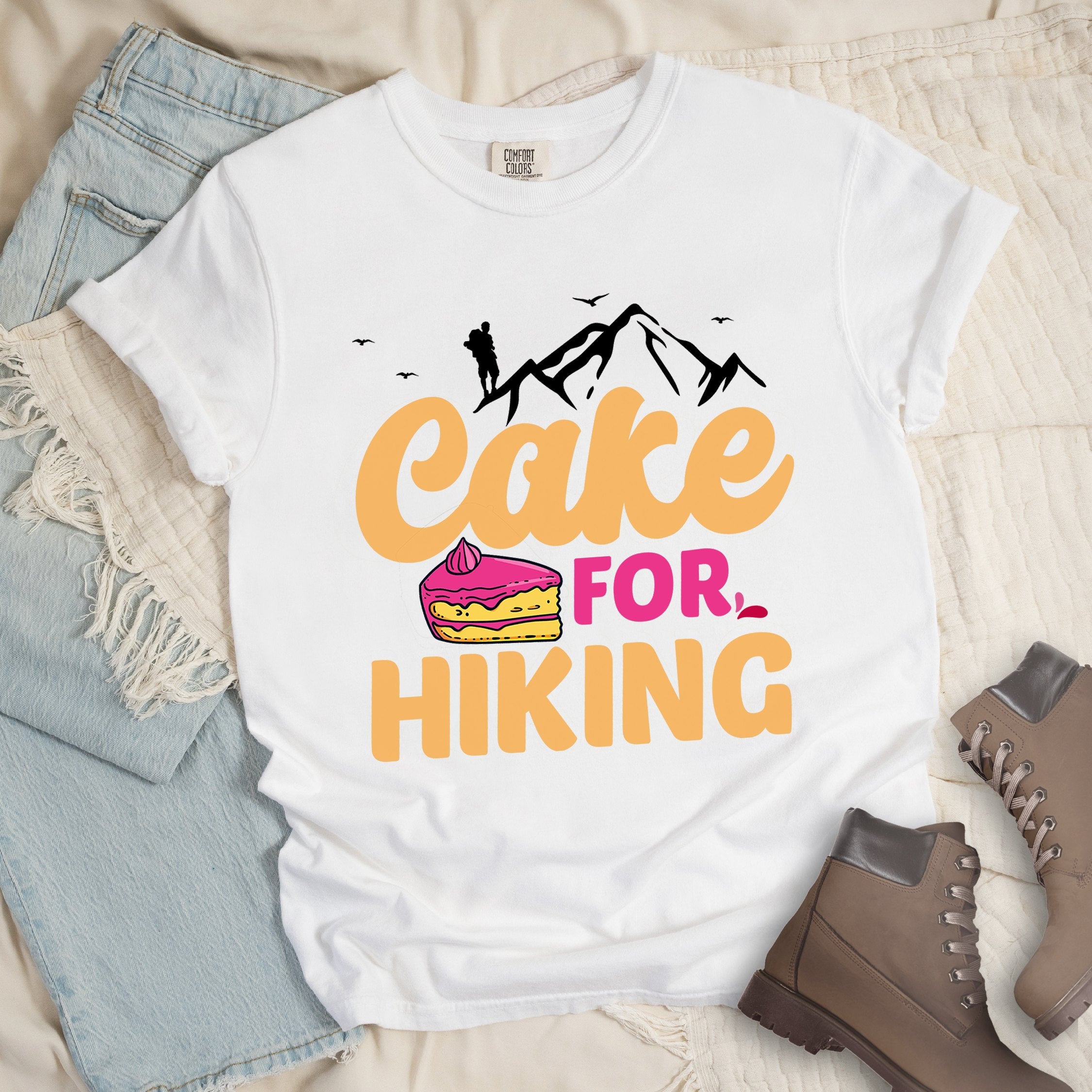White t-shirt with text reading "Cake FOR HIKING". Features white mountain silhouettes with a hiker figure and birds at the top, and an illustration of a yellow cake slice with pink frosting between the words.