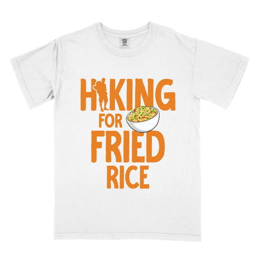 White t-shirt with orange text reading "HIKING FOR FRIED RICE". "HIKING" appears at the top with a hiker silhouette integrated into the letter "I", "FOR" in smaller text in the middle, and "FRIED RICE" in large letters at the bottom. Features an illustration of a white bowl filled with colorful fried rice with vegetables next to "FOR" and above "FRIED".