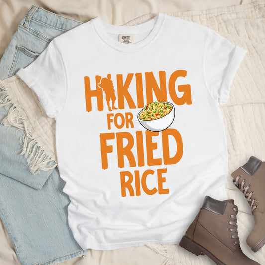 White t-shirt with orange text reading "HIKING FOR FRIED RICE". "HIKING" appears at the top with a hiker silhouette integrated into the letter "I", "FOR" in smaller text in the middle, and "FRIED RICE" in large letters at the bottom. Features an illustration of a white bowl filled with colorful fried rice with vegetables next to "FOR" and above "FRIED". Features a classic crew neck and casual rolled sleeves, styled alongside light wash denim and brown hiking boots. 