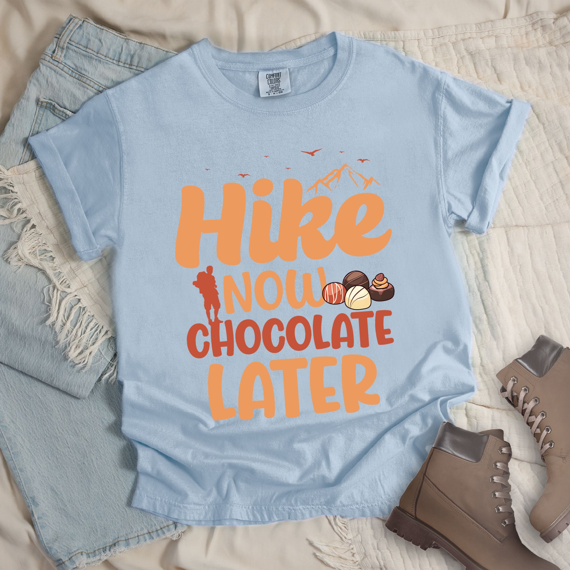Light blue shirt with the phrase “Hike Now Chocolate Later.” Shows a hiker silhouette, mountains, birds, and several chocolate truffles.