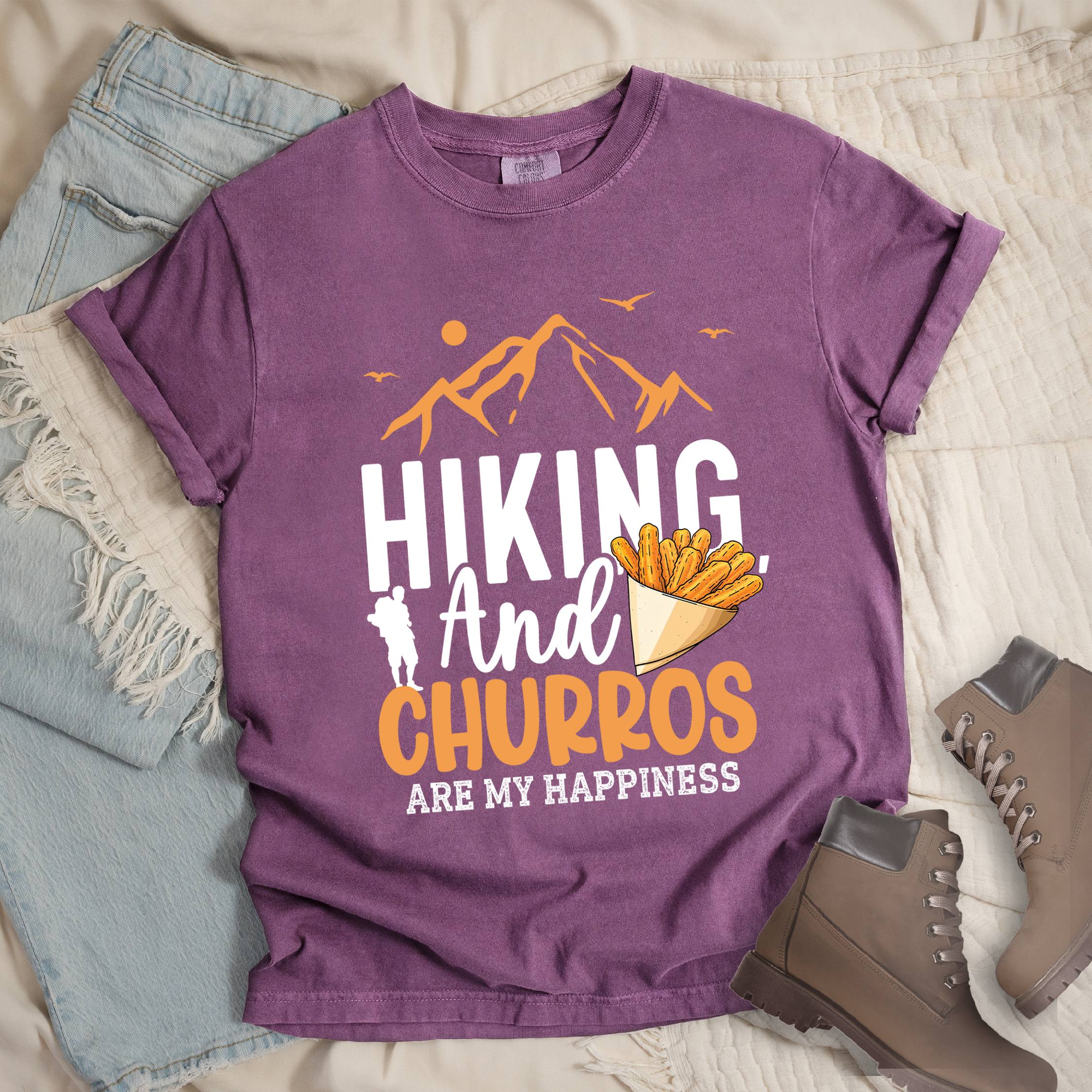 Dark pink shirt with the phrase “Hiking And Churros Are My Happiness.” Features a hiker silhouette, mountains, birds, and a churros cone illustration.