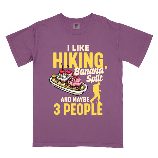 Berry (Dark Violet) t-shirt with yellow and cream text reading "I LIKE HIKING, Banana Split AND MAYBE 3 PEOPLE". "I LIKE" appears in cream at the top, "HIKING," in large yellow letters, "Banana Split" in cream script, "AND MAYBE" in cream, and "3 PEOPLE" in yellow at the bottom. Features a detailed illustration of a banana split dessert with ice cream, cherries and banana slices in the center and a yellow hiker silhouette on the right. Humorously connects hiking, dessert and introversion.