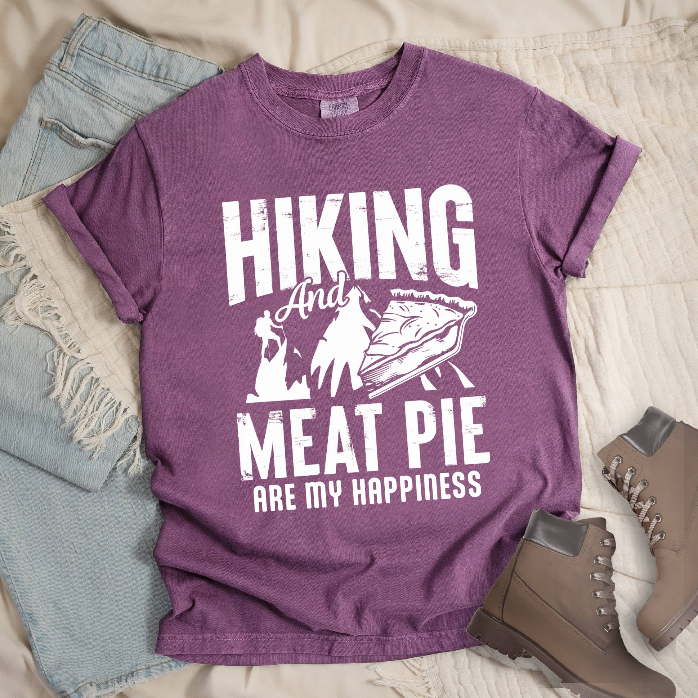 Dark pink shirt showing a hiking design with 'Hiking and meat pie are my happiness' phrase in front of a mountain and hiker silhouette. In front a slice of pie is shown.
