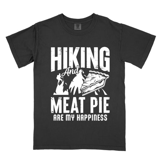 A black t-shirt featuring white distressed text reading "HIKING And MEAT PIE ARE MY HAPPINESS." Between the words "HIKING" and "MEAT PIE" is a graphic showing a mountain silhouette with a tiny hiker climbing it, alongside an illustration of a slice of meat pie. The design uses only white graphics and text on the black shirt background in a rugged, vintage style.