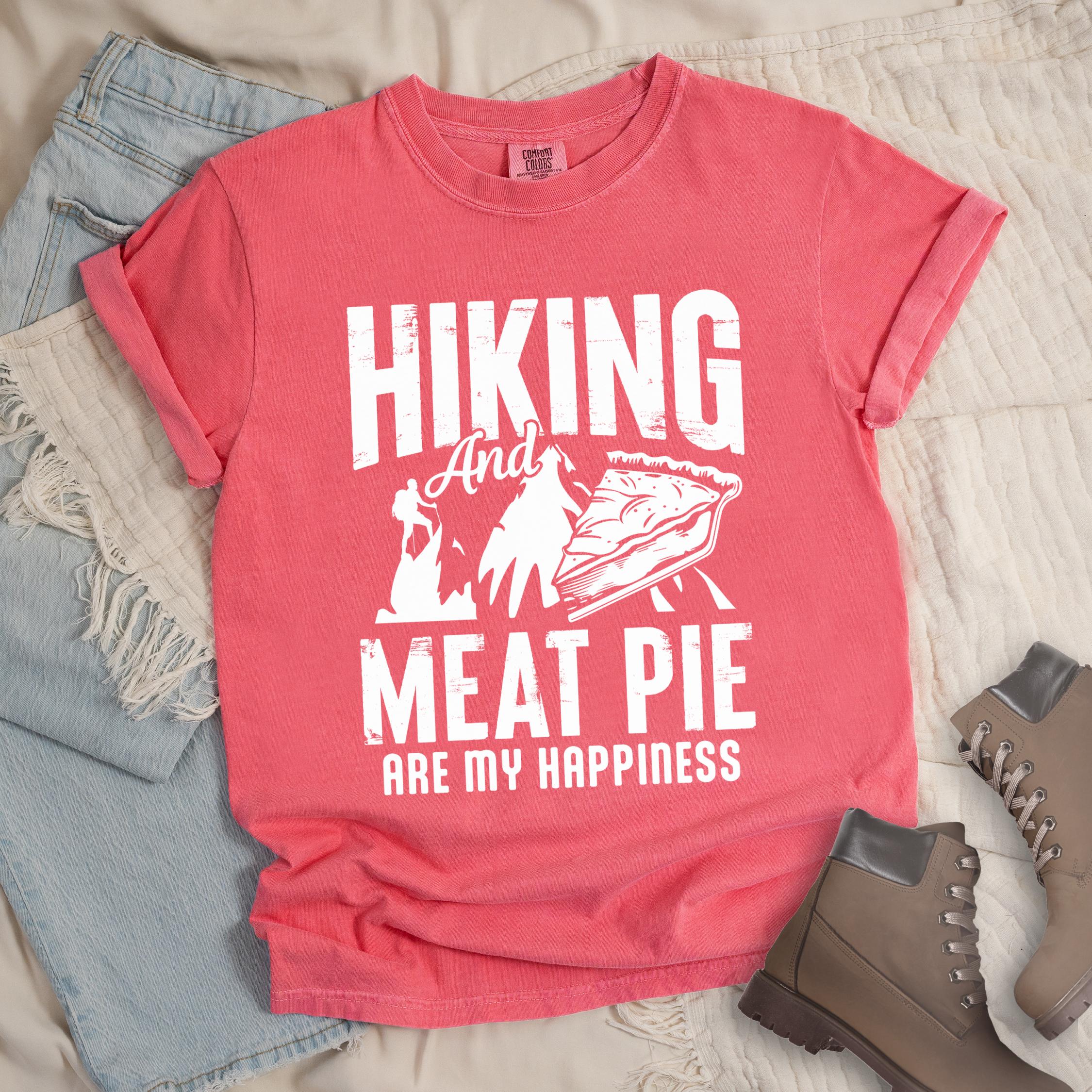 Pink shirt showing a hiking design with 'Hiking and meat pie are my happiness' phrase in front of a mountain and hiker silhouette. In front a slice of pie is shown.