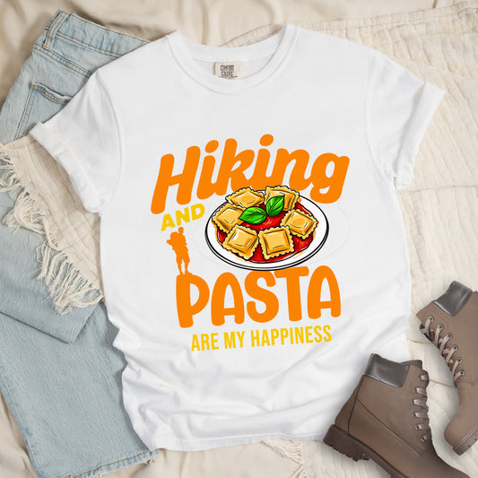 White t-shirt with text "Hiking AND PASTA ARE MY HAPPINESS". "Hiking" appears in stylized orange text at the top, "AND" in smaller yellow text, "PASTA" in large orange letters, and "ARE MY HAPPINESS" in small yellow text at the bottom. Features an orange silhouette of a hiker with backpack on the left and an illustration of ravioli pasta with tomato sauce and basil leaves on a white plate. Features a classic crew neck and casual rolled sleeves, styled alongside light wash denim and brown hiking boots. 