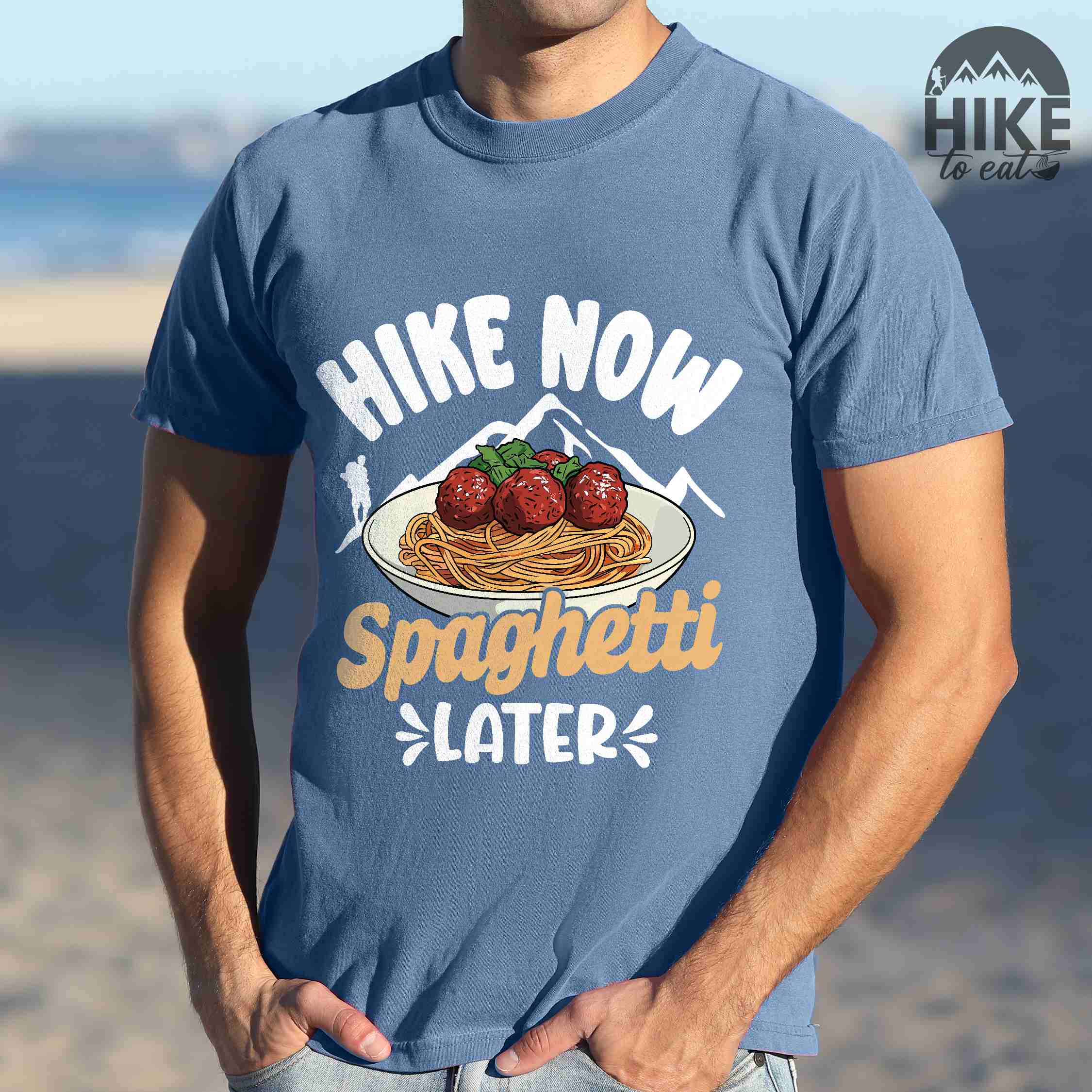 Man wearing blue jean "Hike now spaghetti later" colored vintage tee with bold typography, mountains, birds, illustrated plate of spaghetti meatballs, and backpackers for pasta lovers.